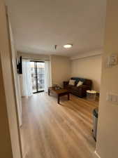 Bright living area featuring wide-plank wood-finish flooring, a wall-mounted flat-screen television, a full-light glass door opening to a balcony, and a ceiling-mounted light fixture