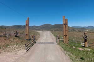 Gated property entrance featuring a dirt road, wooden posts with wagon wheel accents, and a metal fence