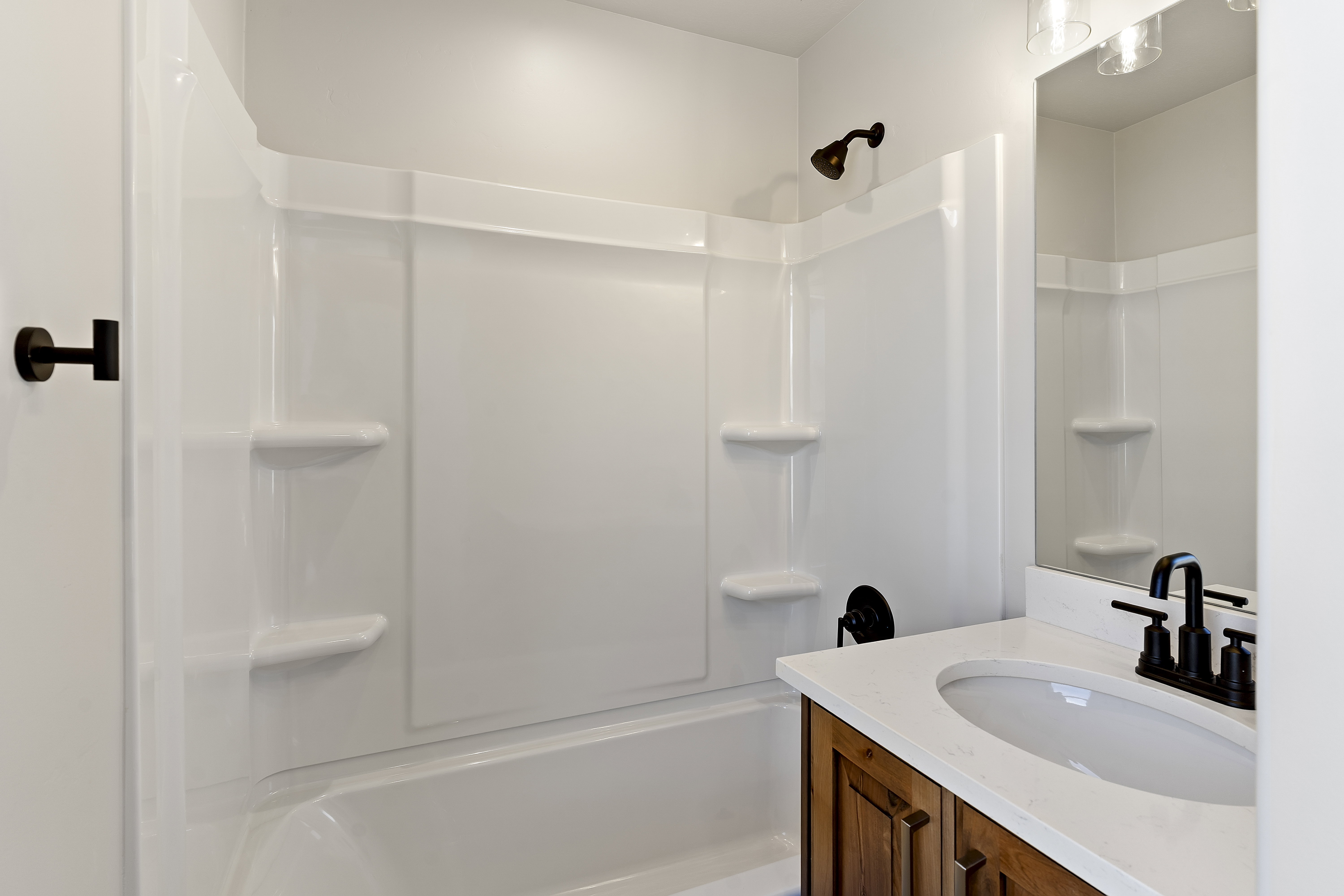 Bathroom featuring a white tub and shower combination with integrated shelving, a wood-finish vanity with panel detailing, a white countertop with an undermount sink, and matte black fixtures