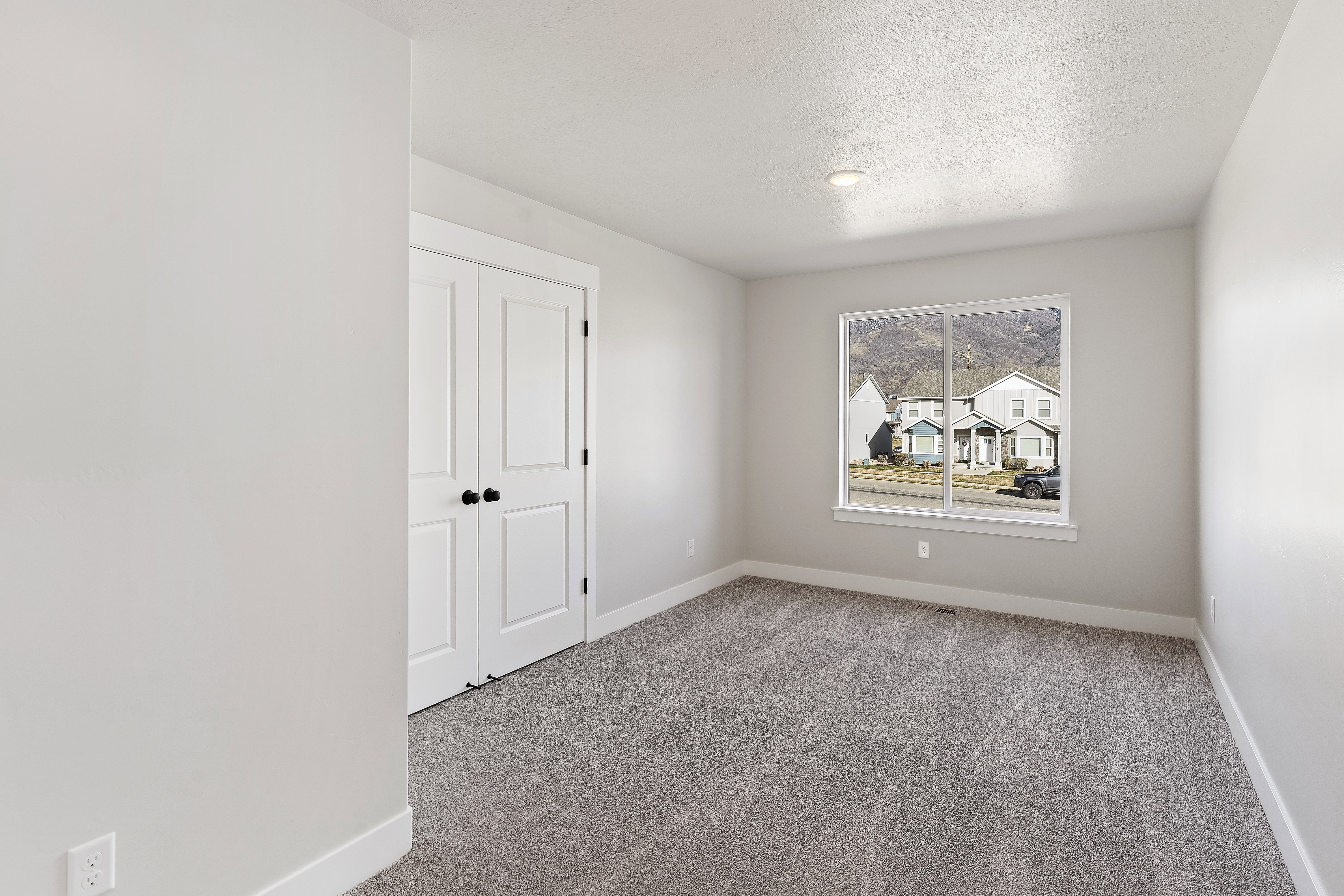 Spacious room featuring neutral carpeting, a large window, white baseboards, and double closet doors with black hardware