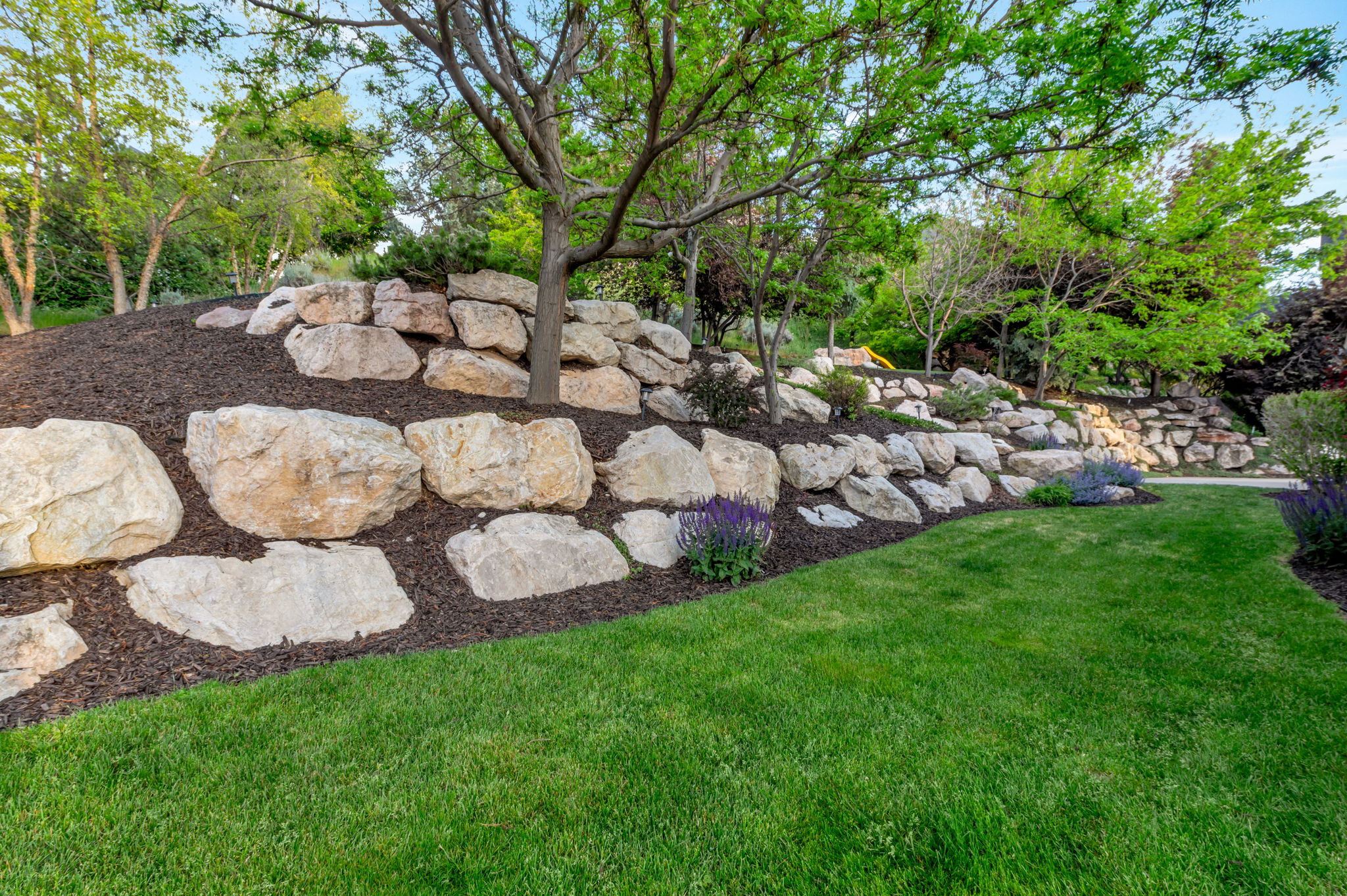 Professionally designed landscaping showcases impressive natural boulder retaining walls, mature trees, and lush green lawn.
