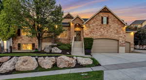 Stone and brick facade featuring an arched entryway with a wooden door, a multi-car garage with arched doors, mature trees, and a landscaped rock garden