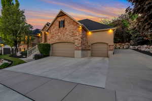 Stone and stucco exterior featuring multiple garages, a concrete driveway, and a tiered landscaped yard with mature trees