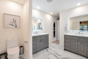 Spacious bathroom featuring dual gray vanities with white countertops, illuminated mirrors, recessed lighting, and large format marble-look tile flooring
