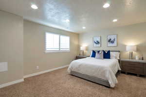 Recessed ceiling lighting illuminates a spacious room featuring textured carpet flooring, neutral-toned walls, and a window with white plantation shutters