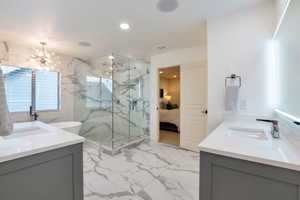 Spacious bathroom featuring large format marble-look tile flooring and walls, a glass-enclosed shower, dual vanities with white countertops, and a freestanding soaking tub