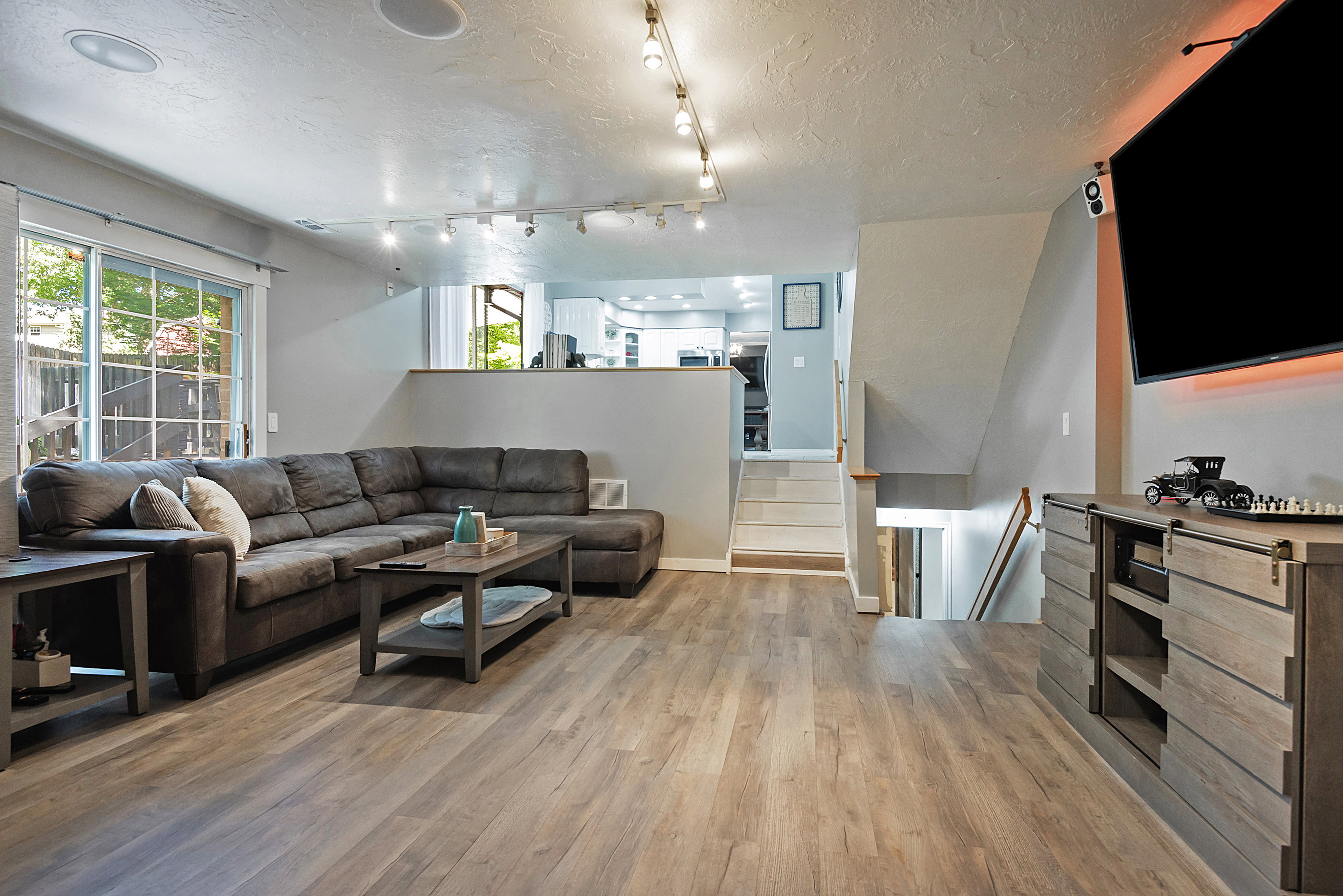 Spacious main living area featuring wood-finish flooring, a patio door with exterior views, track lighting, recessed ceiling speakers, and a half-wall opening to an upper level