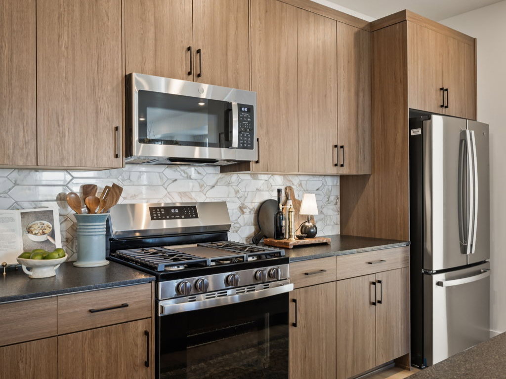 Contemporary kitchen featuring wood-finish cabinetry, stainless steel appliances, dark countertops, a patterned tile backsplash, and recessed lighting