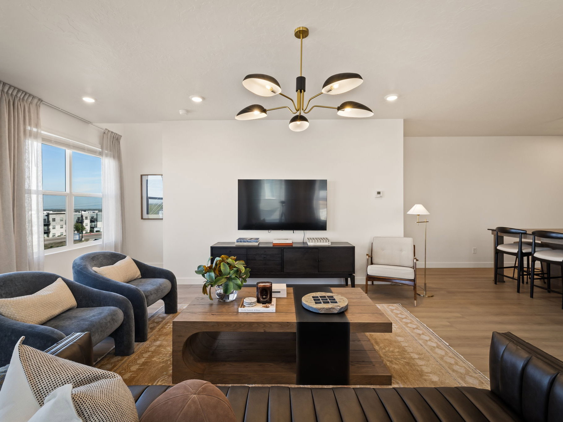 Spacious open-concept living area featuring light wood-finish flooring, a contemporary chandelier, recessed lighting, and large windows with abundant natural light