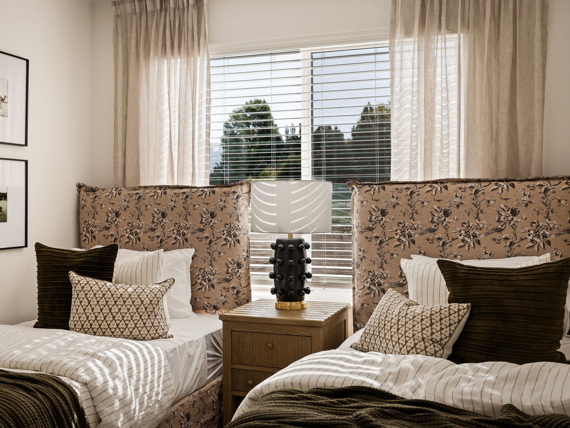 Room featuring two beds with patterned upholstered headboards, a central window with horizontal blinds and sheer curtains, and a wooden nightstand