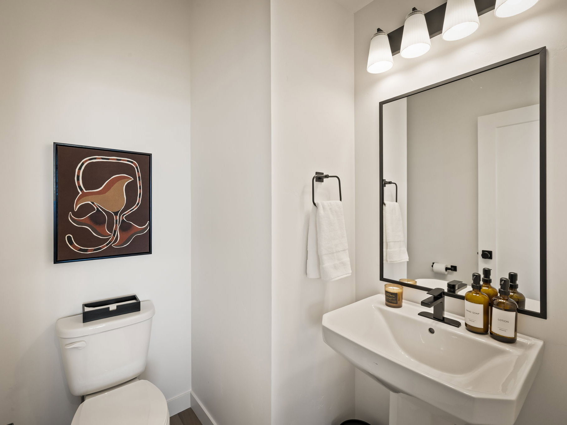 Powder room featuring a pedestal sink with matte black faucet, wall-mounted rectangular mirror with black frame, and a four-light vanity fixture