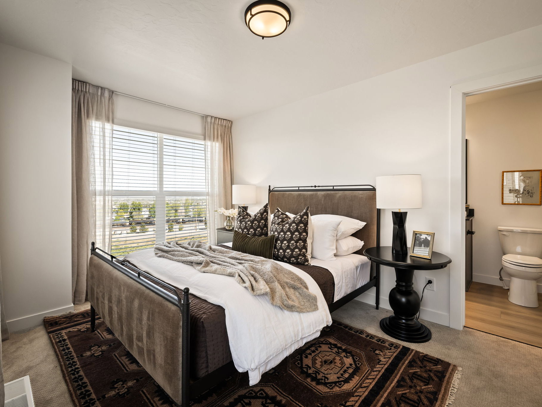 Carpeted bedroom featuring a large window with blinds and drapes, an overhead flush-mount light fixture, and an en-suite bathroom with wood-finish flooring