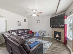 Living area featuring wood-finish flooring, a corner fireplace with a tiled surround, a ceiling fan, and a large window with blinds