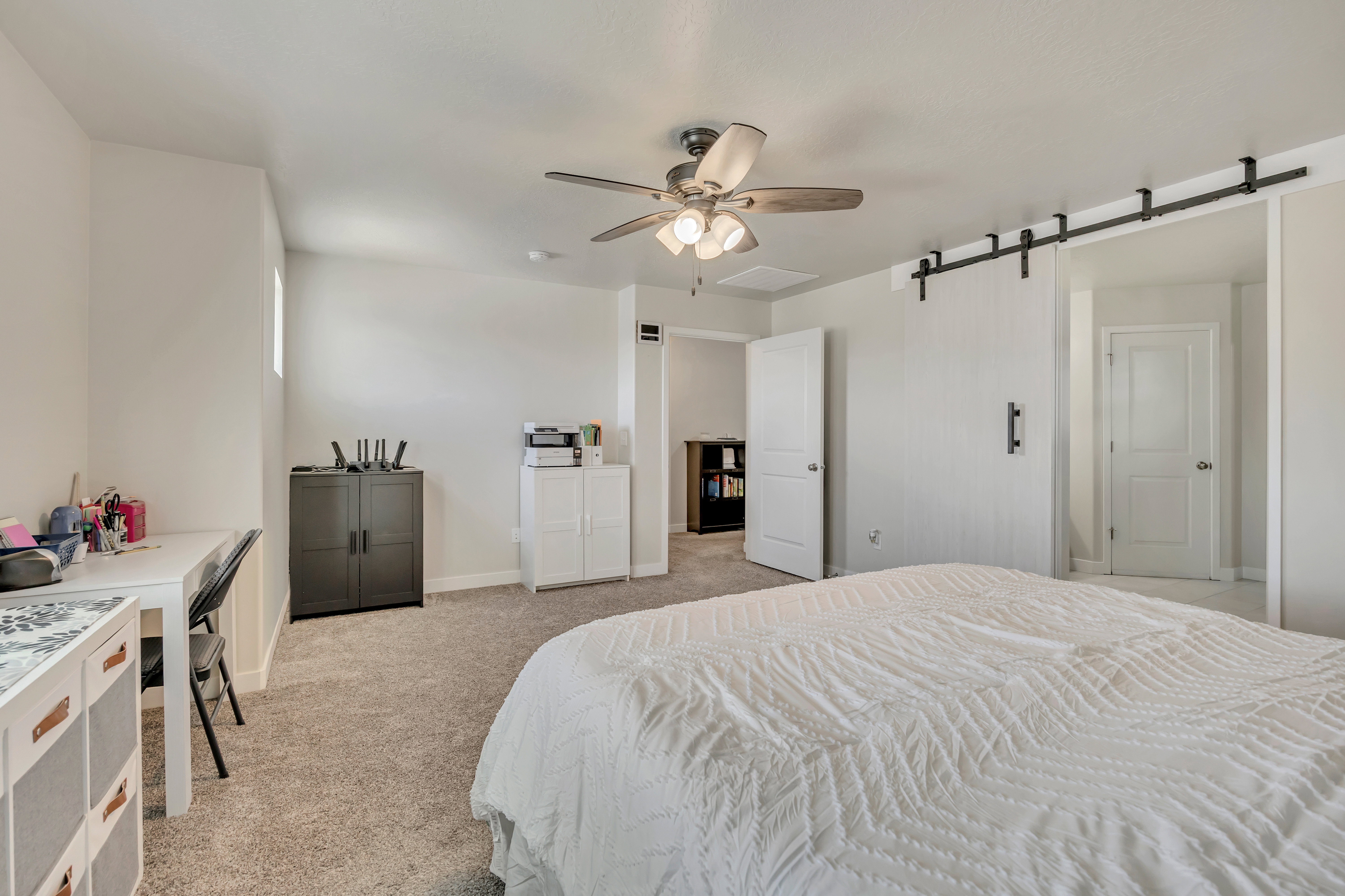 Carpeted room featuring a ceiling fan with light fixture, a window providing natural light, and a modern barn door on a black metal track