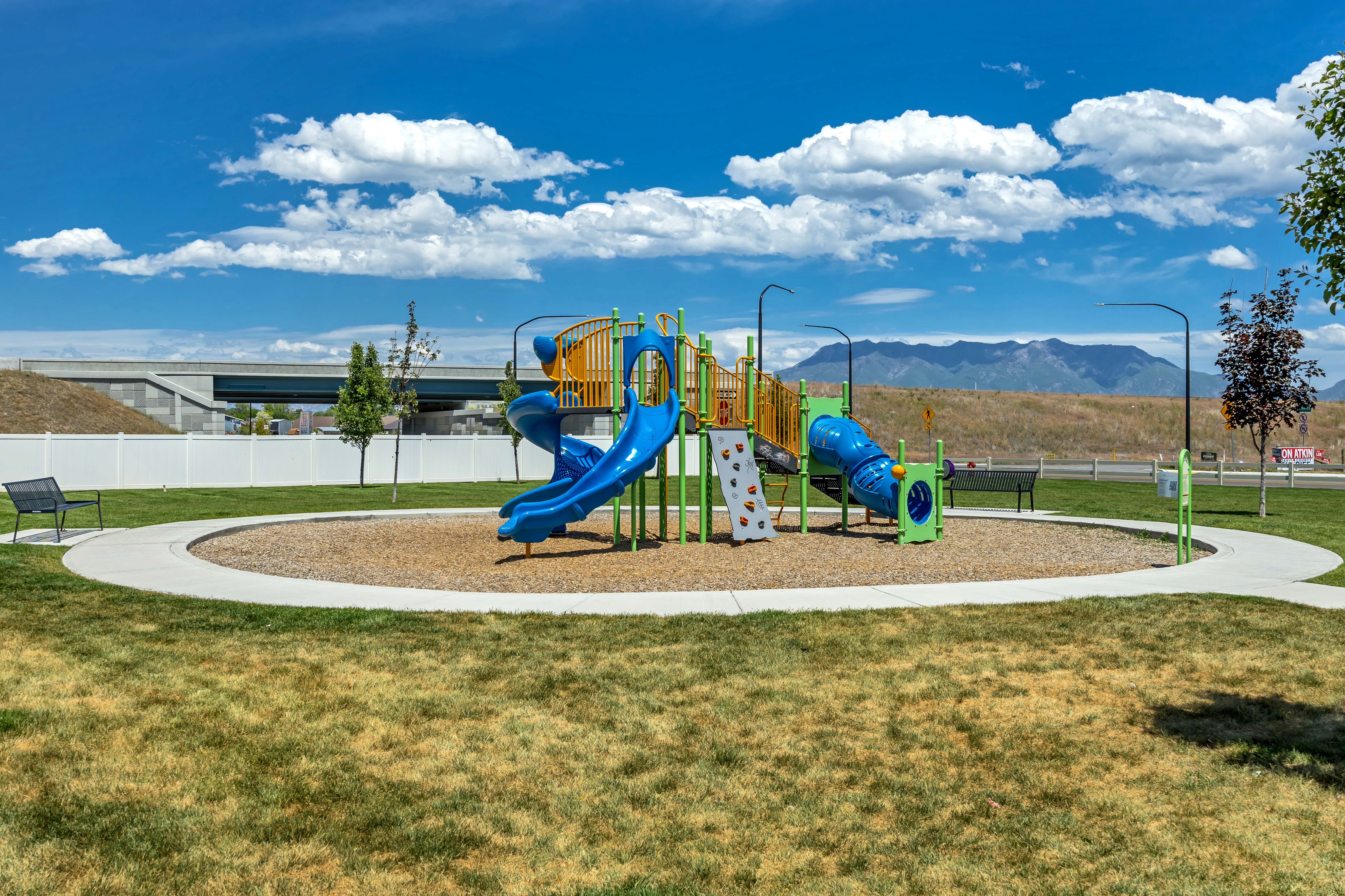 Community playground featuring a multi-level play structure with slides and climbing walls, surrounded by a wood chip surface and a concrete walking path