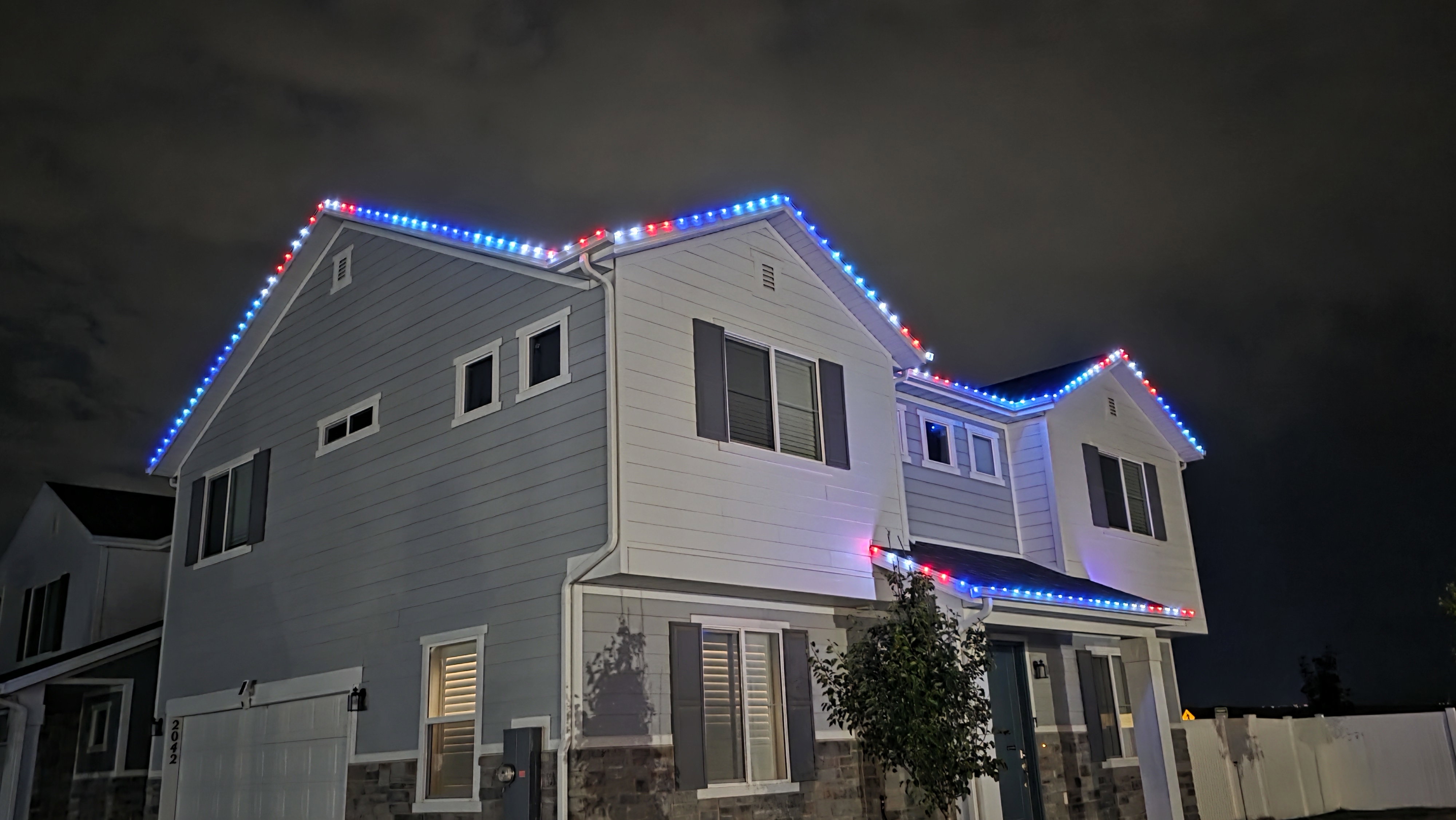 Two-story residence featuring light-toned siding and dark shutters-permanent exterior custom holiday lighting