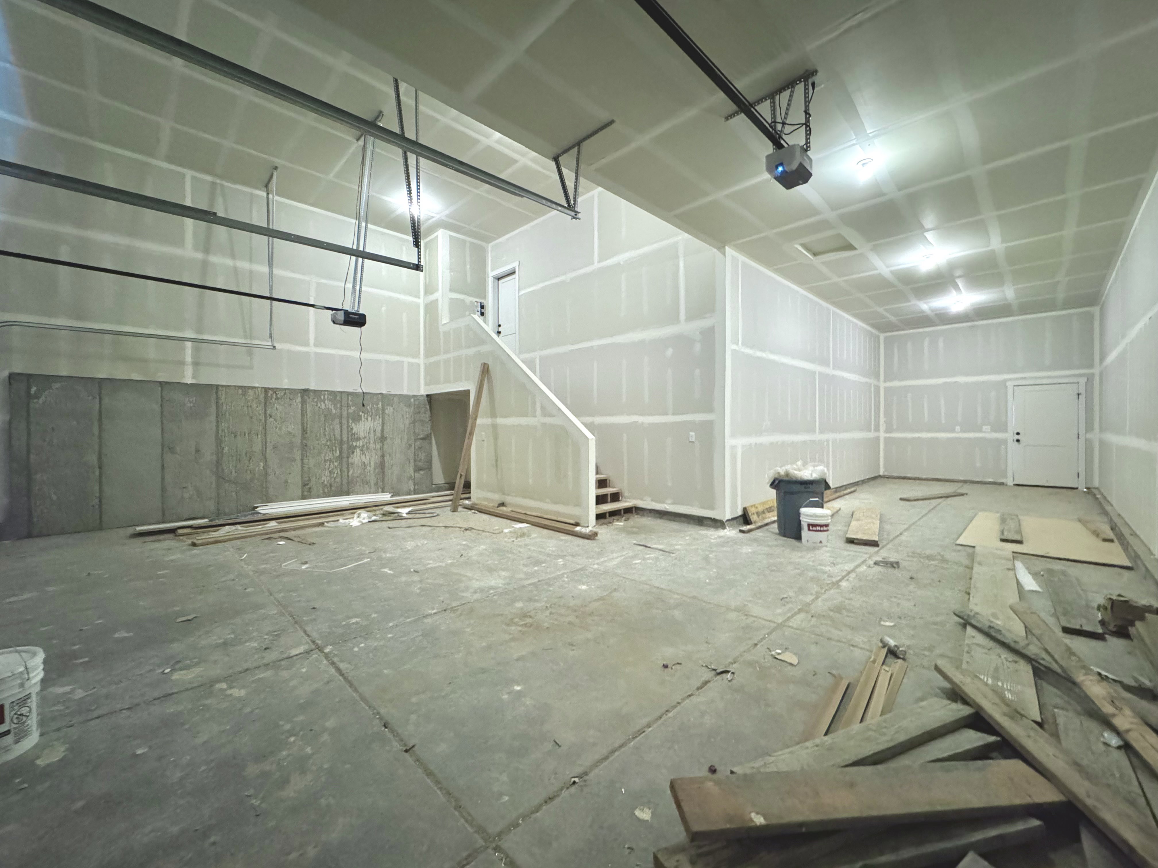 Expansive interior space featuring unfinished drywall throughout, exposed concrete flooring, a partial staircase, and a visible garage door opener mechanism