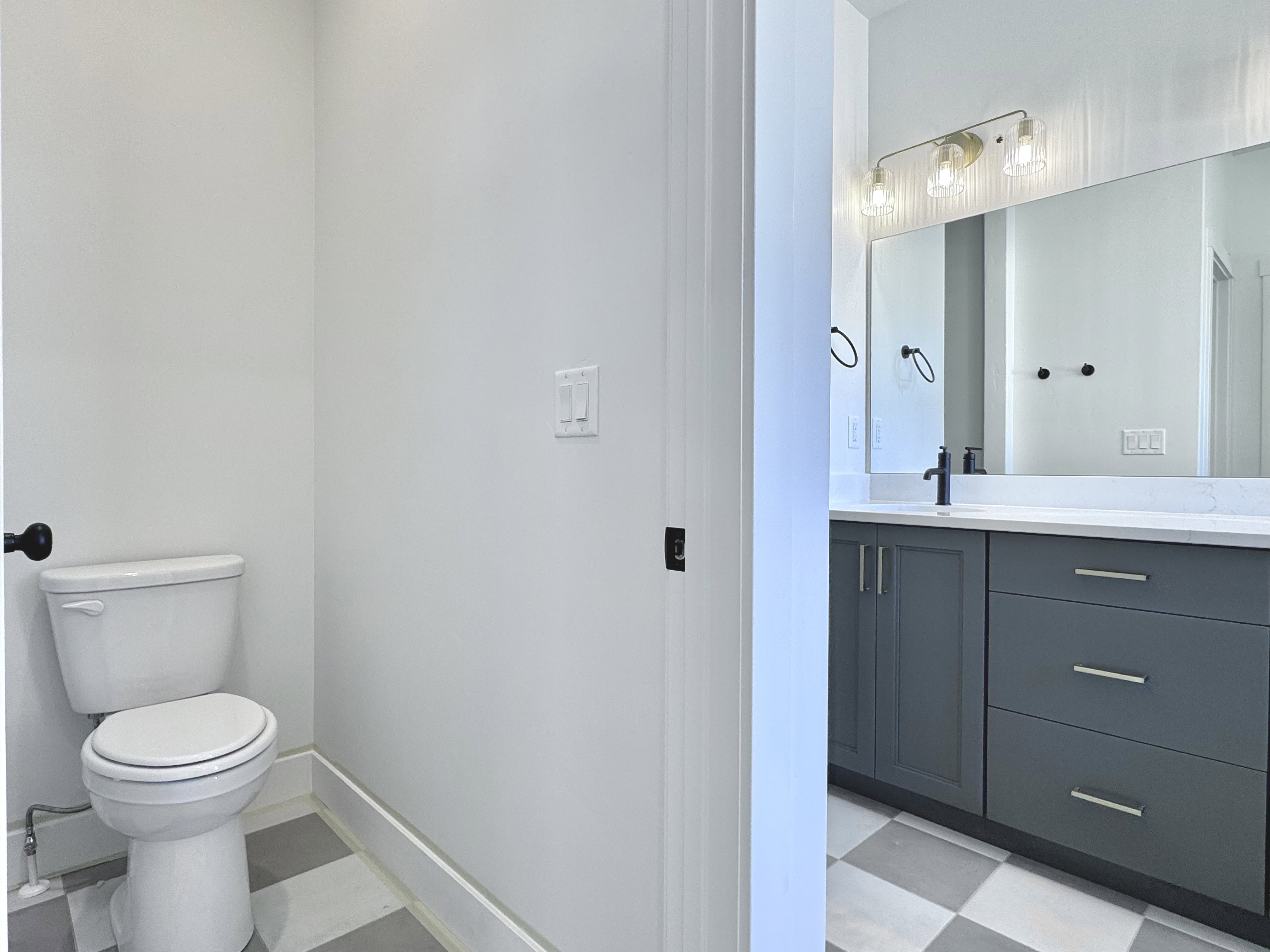 Bathroom featuring a white toilet, checkerboard floor tiles, dark gray vanity with chrome hardware, white countertop, and a large mirror with a three-light vanity fixture