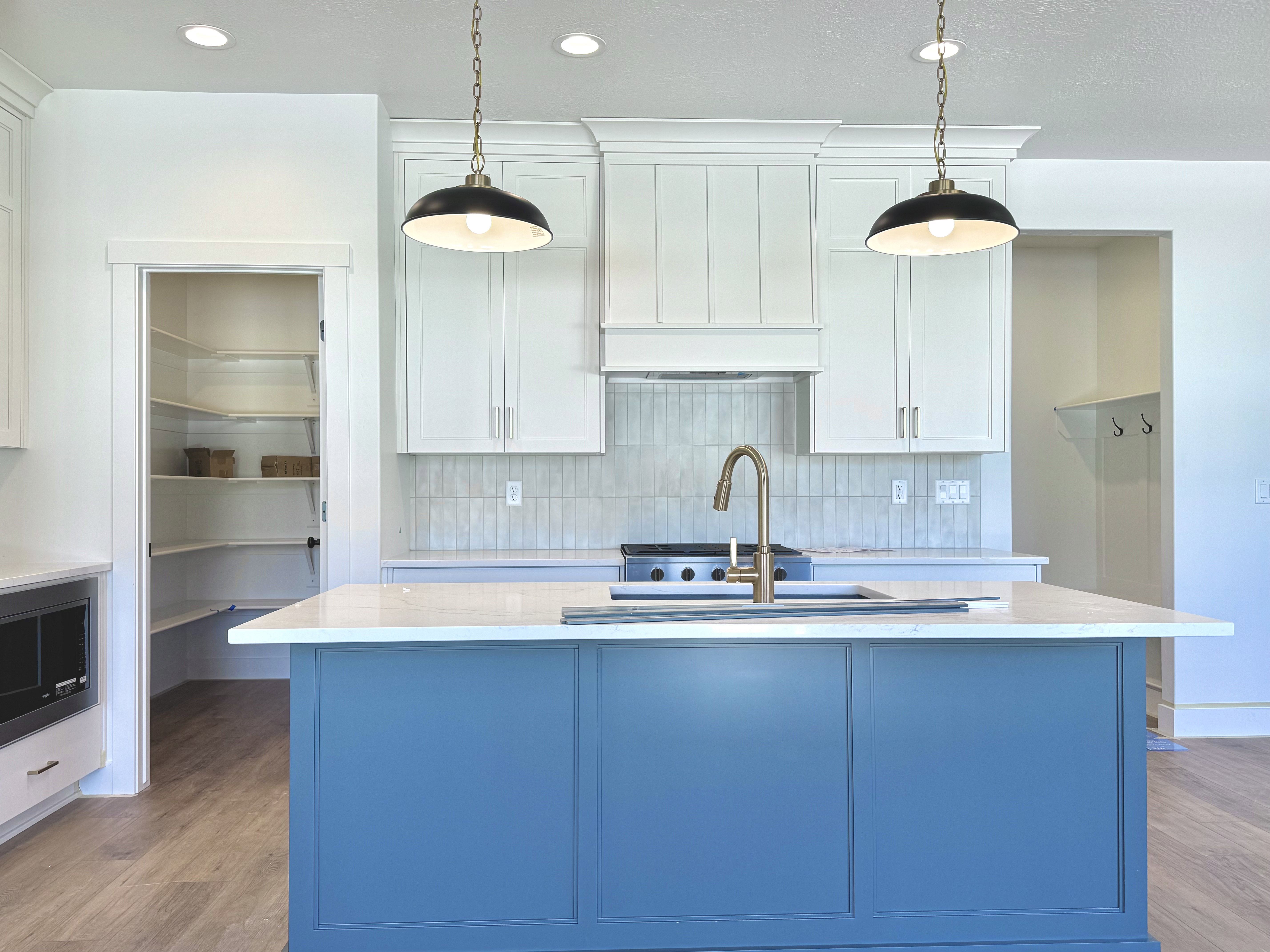 Kitchen featuring a blue island with a built-in sink and brass faucet, white shaker cabinetry, an integrated range hood, white tile backsplash, and recessed lighting