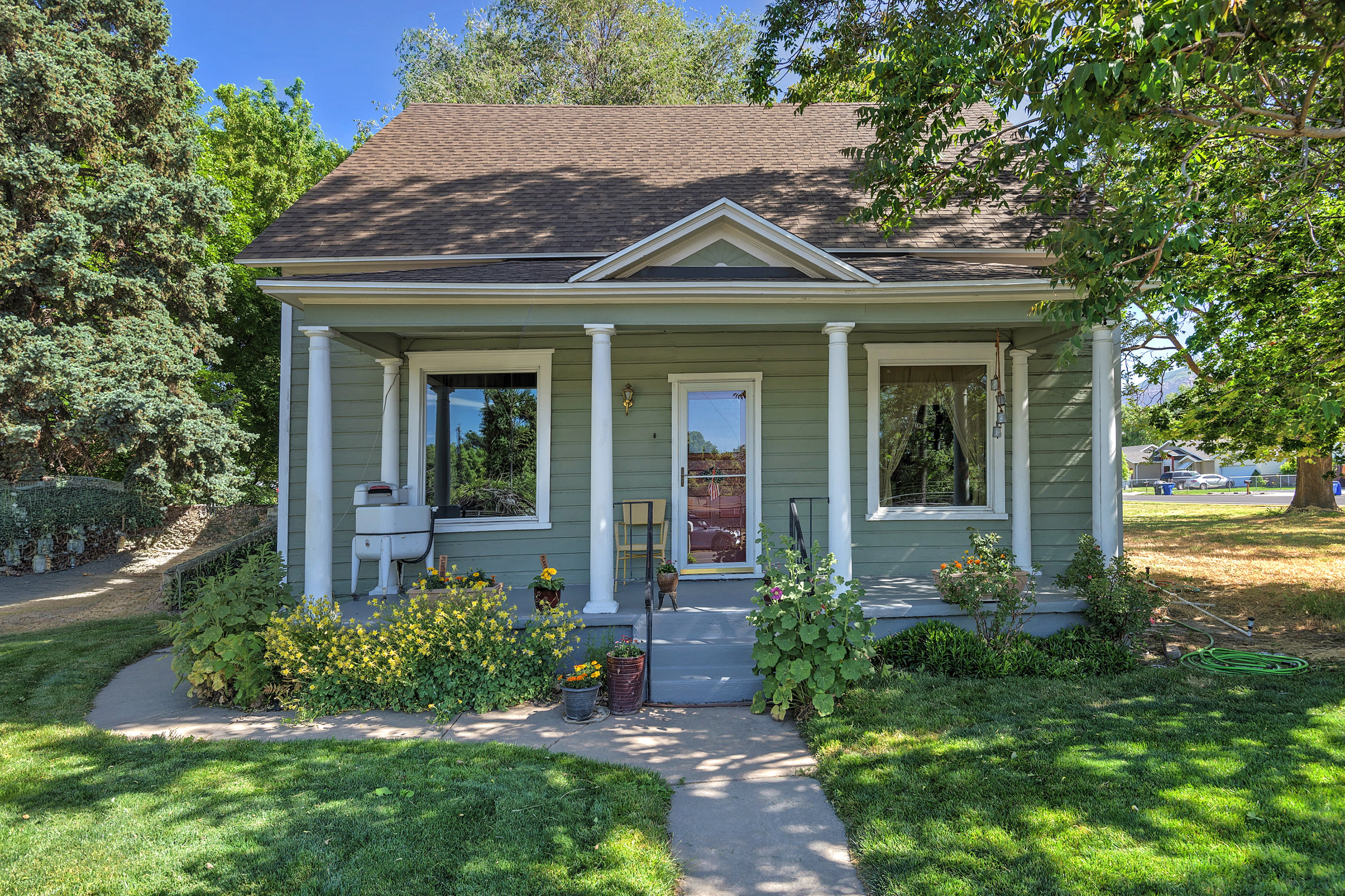 Green exterior with white trim featuring a covered porch with white columns, a dark shingle roof, and a mature tree providing shade