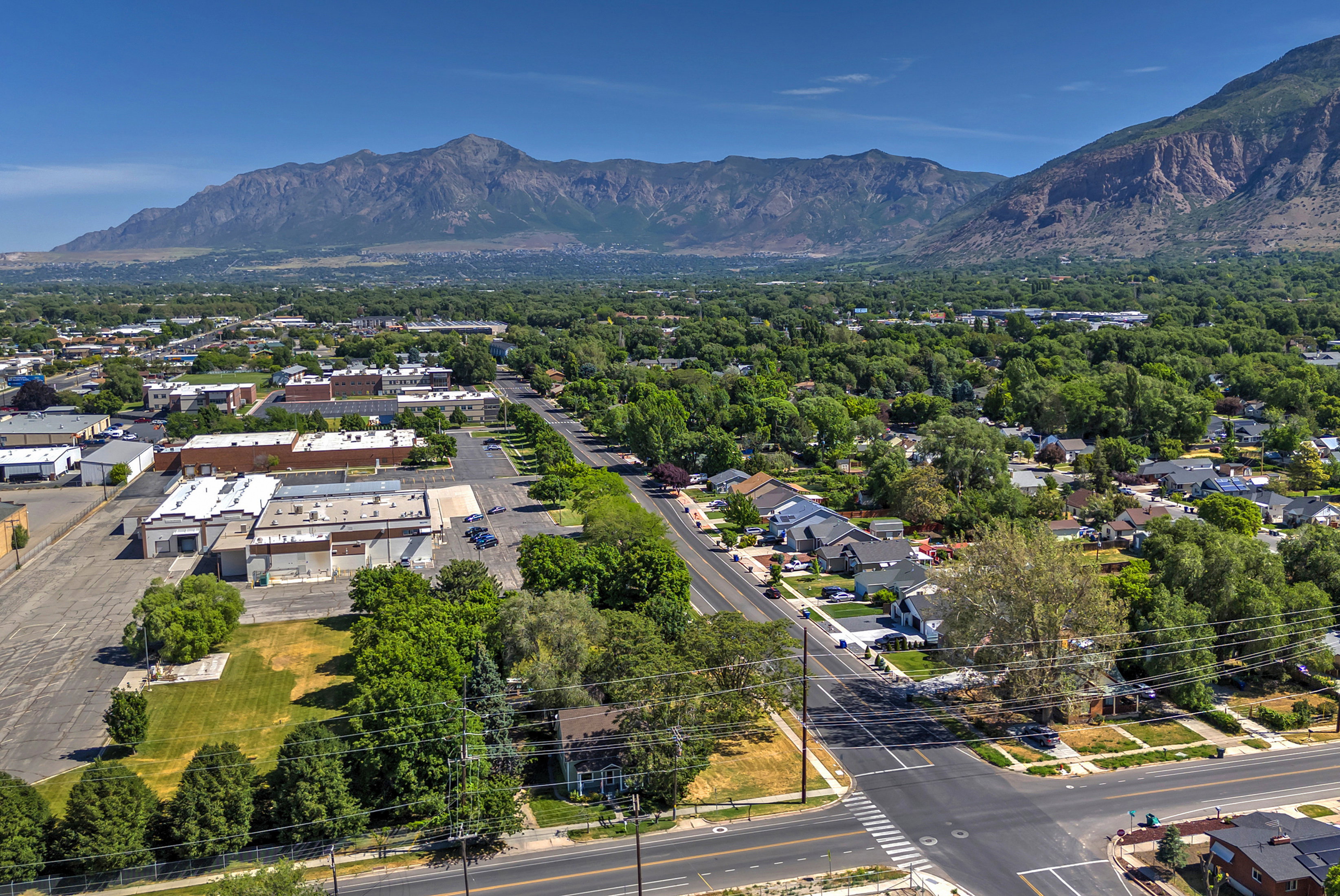 Expansive aerial perspective showcasing a residential neighborhood with tree-lined streets, a commercial area, and a prominent mountain range backdrop