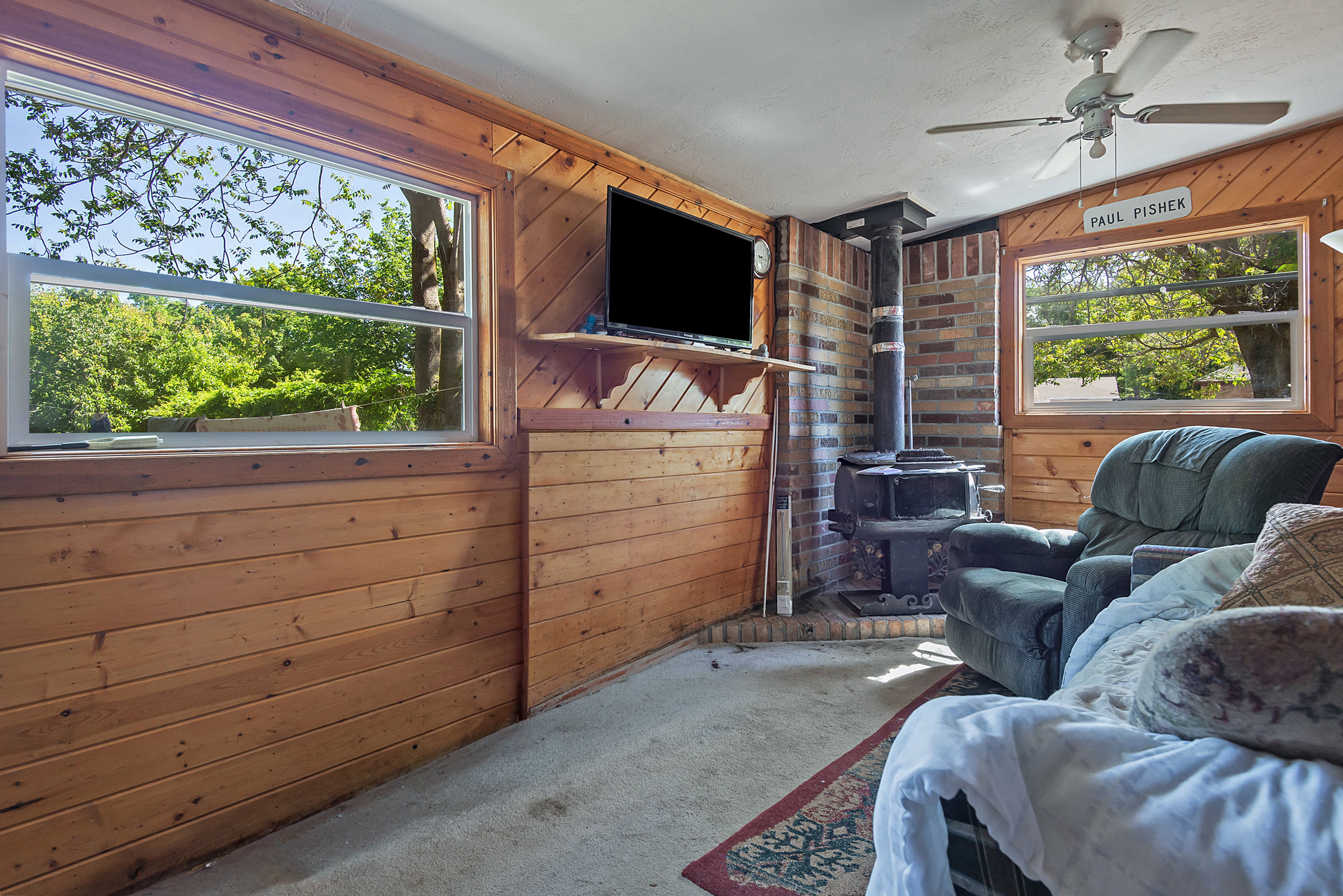 Room featuring wood paneling, a ceiling fan, and a freestanding wood-burning stove with a brick surround
