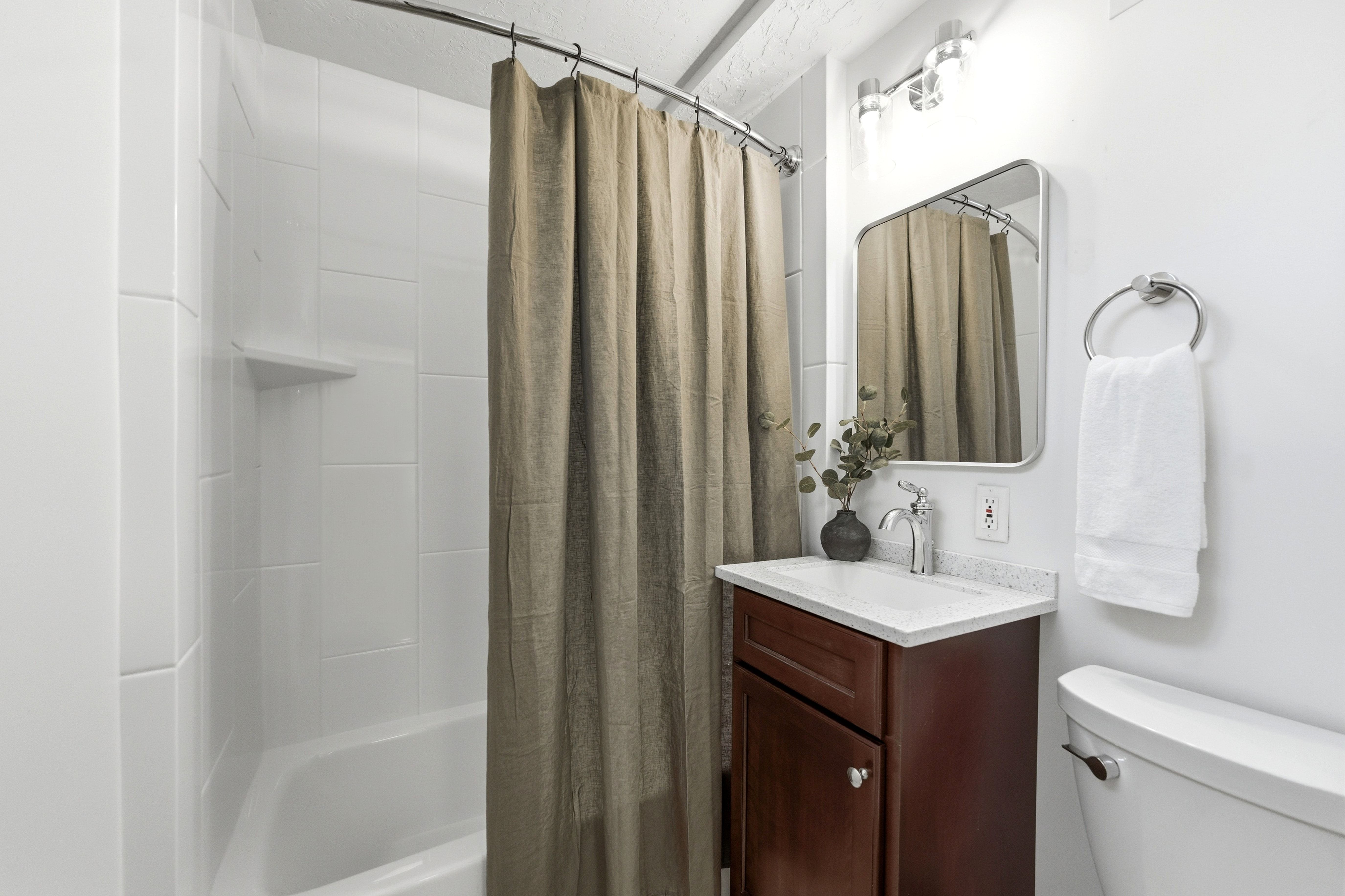 Full bathroom featuring a white tile shower surround, a white bathtub, a dark wood-finish vanity with a white countertop, a rectangular mirror, and a chrome light fixture