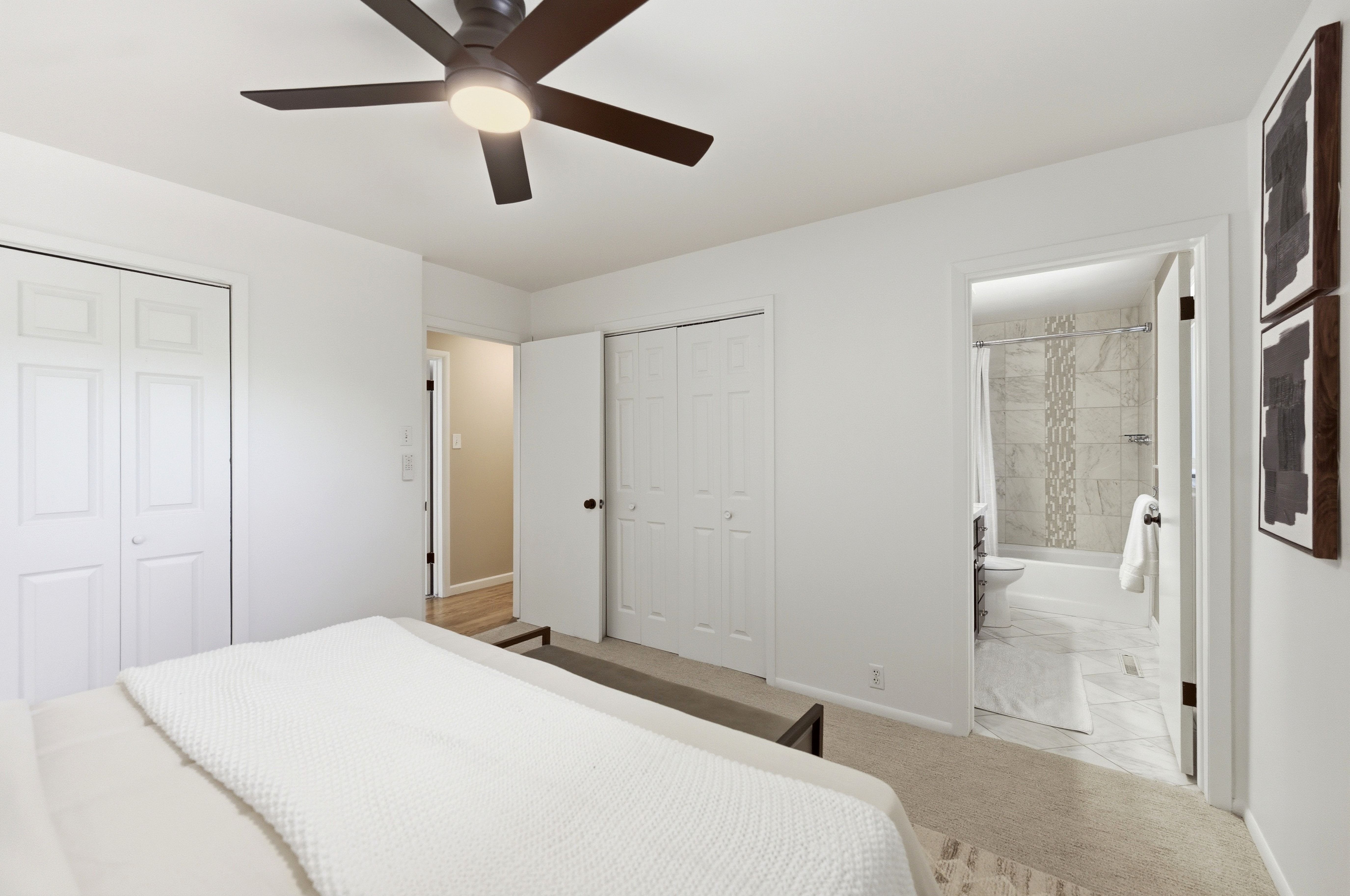 Neutral colored bedroom featuring a ceiling fan with integrated lighting, two bi-fold closet doors, and an en-suite bathroom with tiled flooring and a tiled shower surround