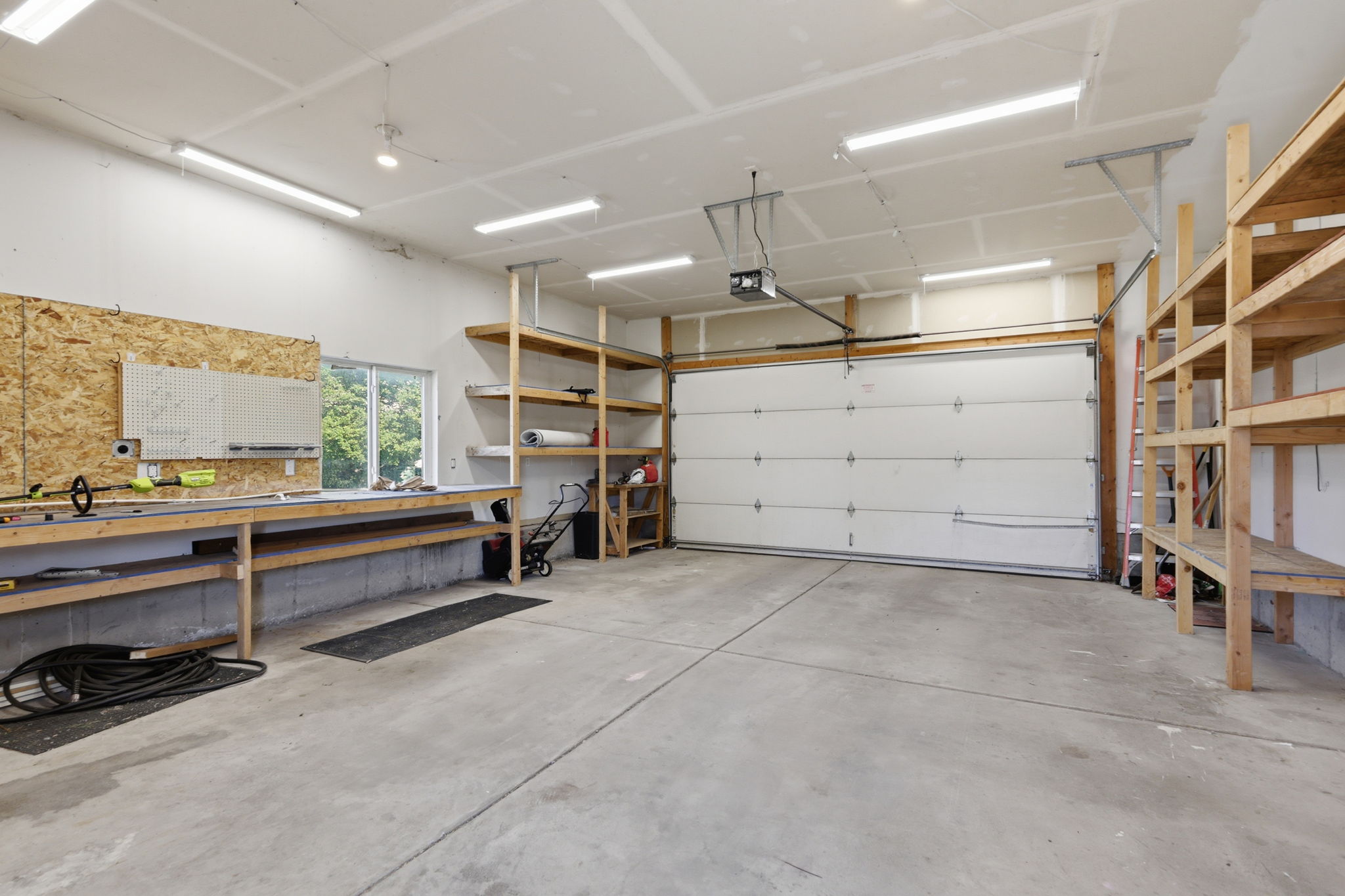 Spacious garage featuring a concrete floor, built-in wooden shelving, a dedicated workbench with an oriented strand board backing, and ample overhead lighting