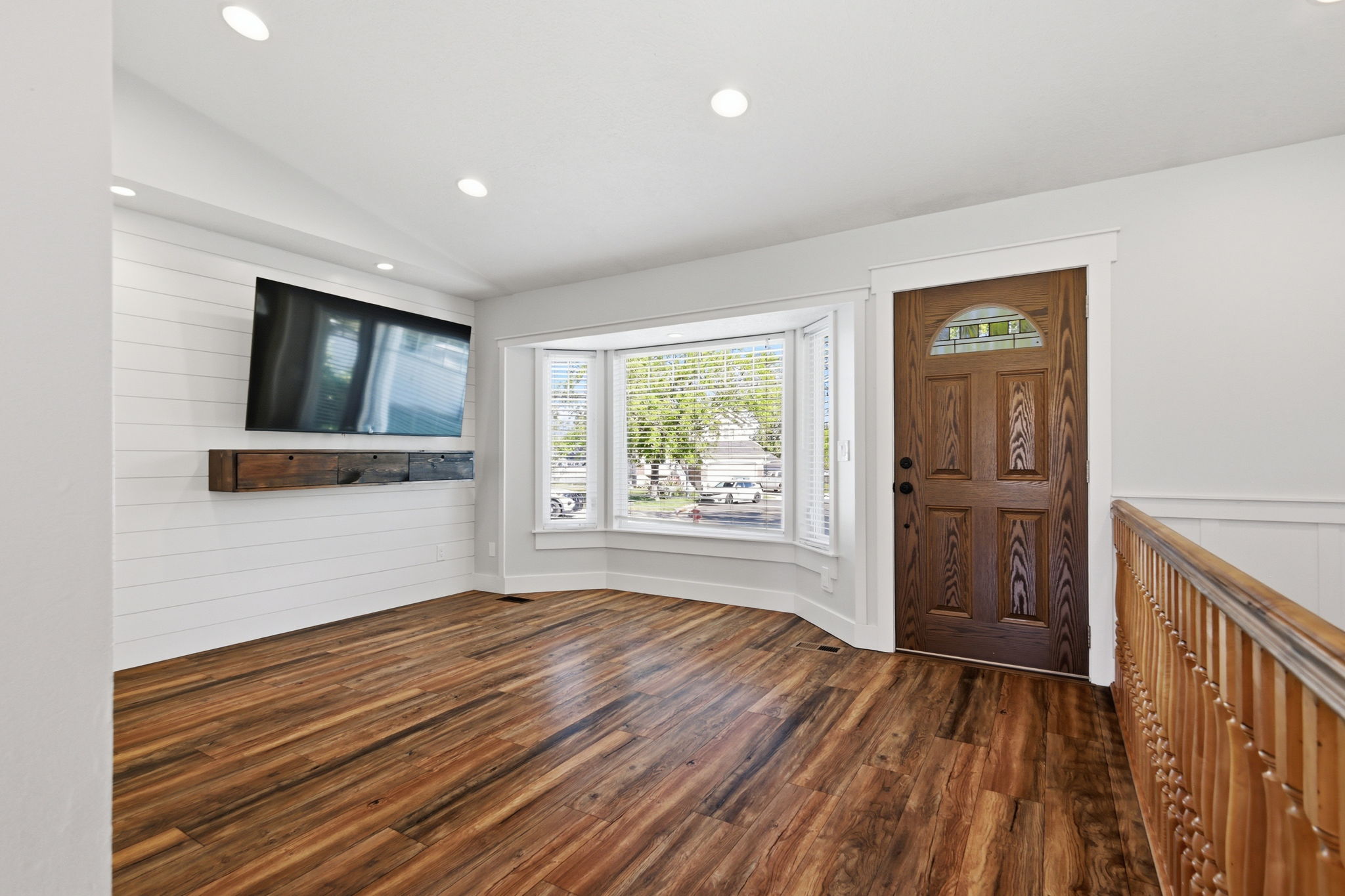 Spacious room featuring wood-finish flooring, a decorative paneled entry door with an arched glass insert, and a bay window with blinds