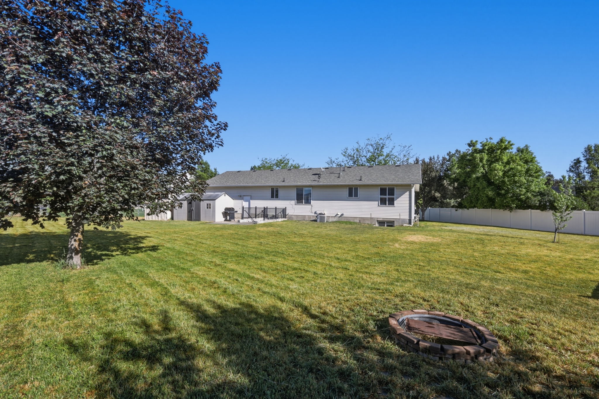 Expansive grassy backyard featuring a large mature tree, a paved deck, a built-in fire pit, and a white privacy fence