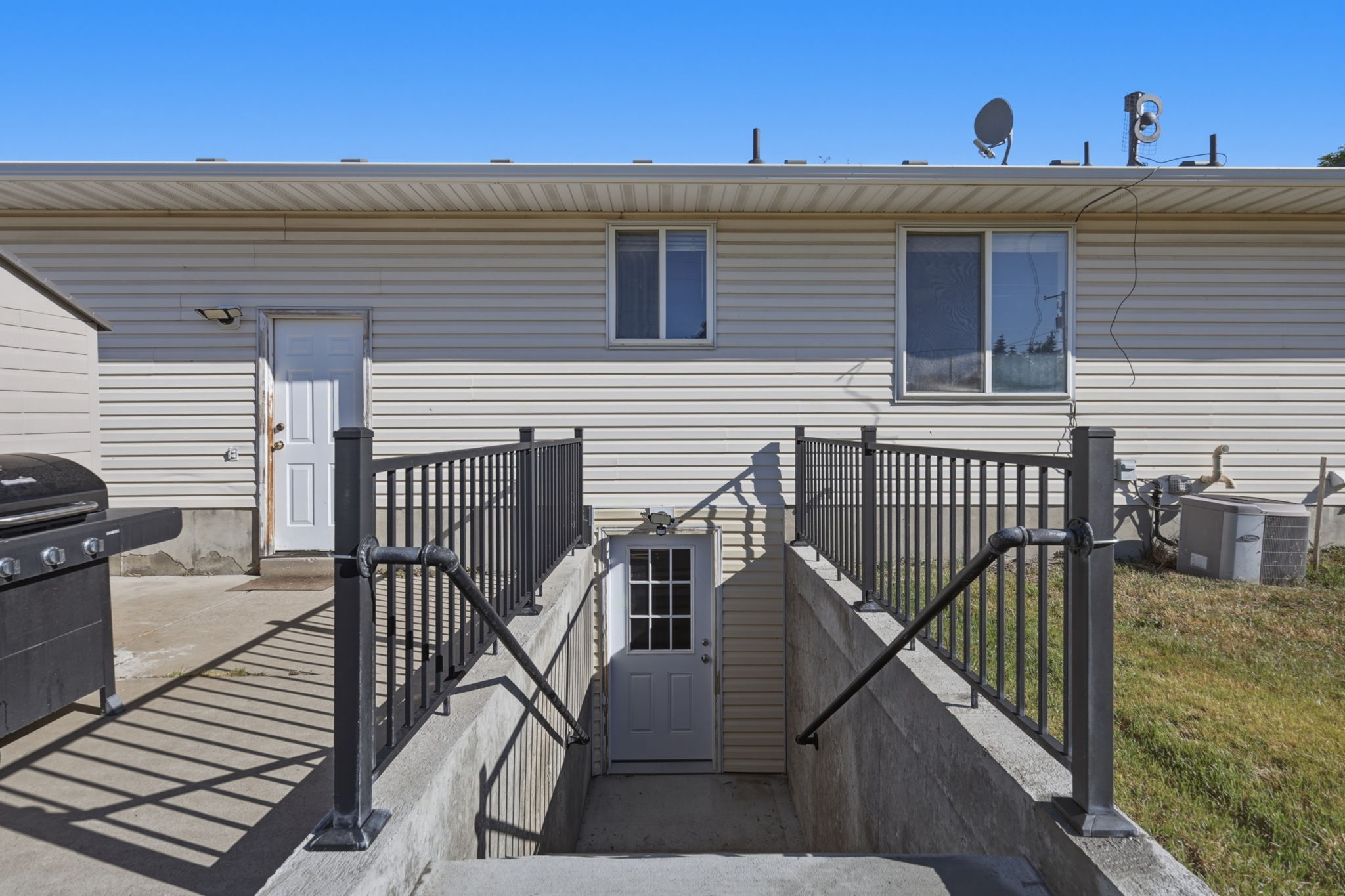 Exterior with light-colored siding, featuring a lower-level concrete stairwell entry with black metal railings
