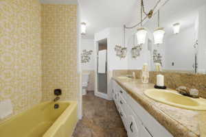 Bathroom featuring a yellow bathtub, yellow patterned wall covering, a dual vanity with a patterned countertop, dual yellow sinks, and a built-in shower enclosure