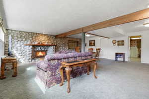 Stone-surround fireplace with a wood mantle, exposed wood beam ceiling, textured white ceilings, and light-toned carpet flooring
