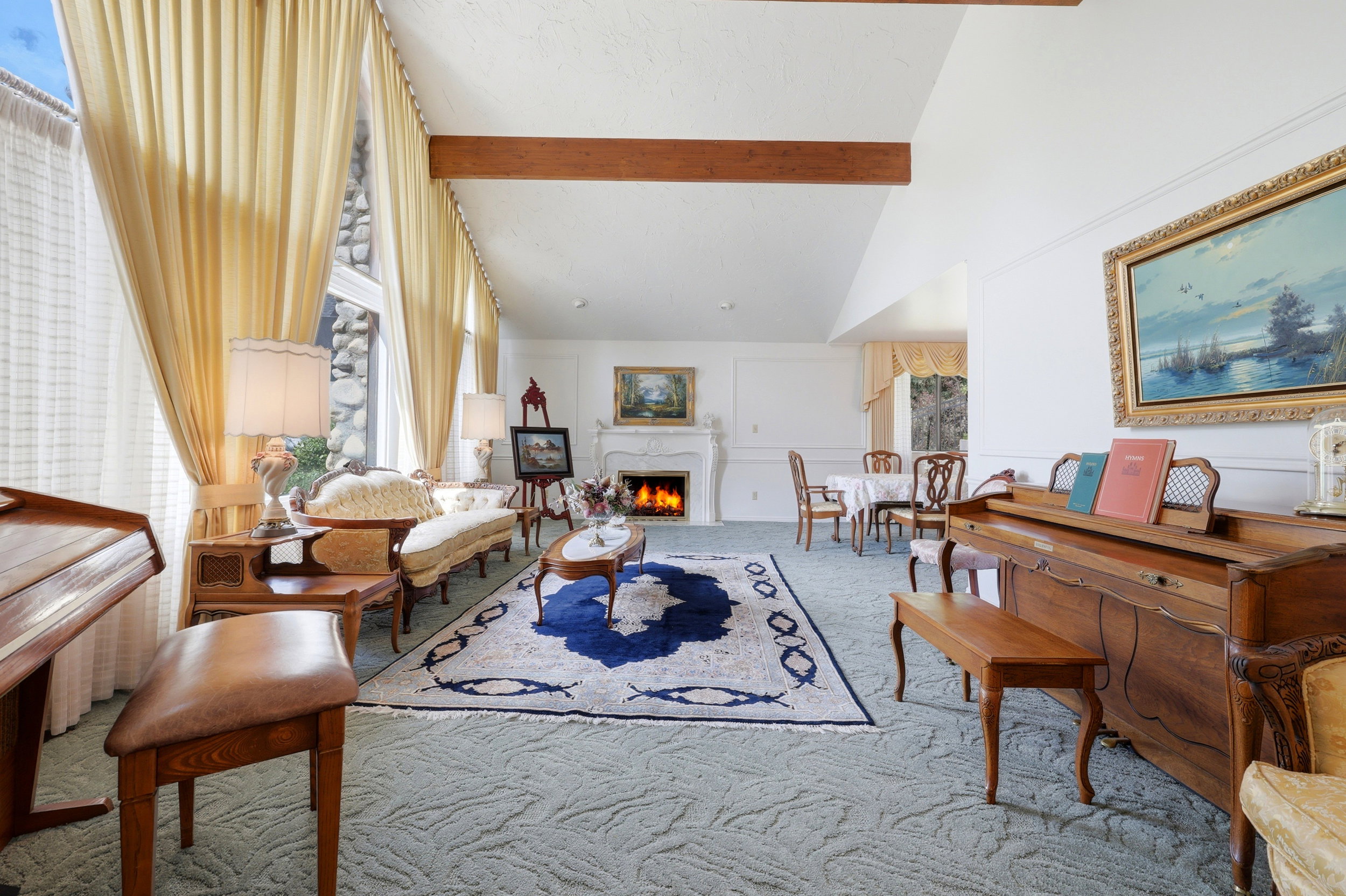 Spacious living area featuring exposed wood beams, a white mantel fireplace, and a large window with a stone surround
