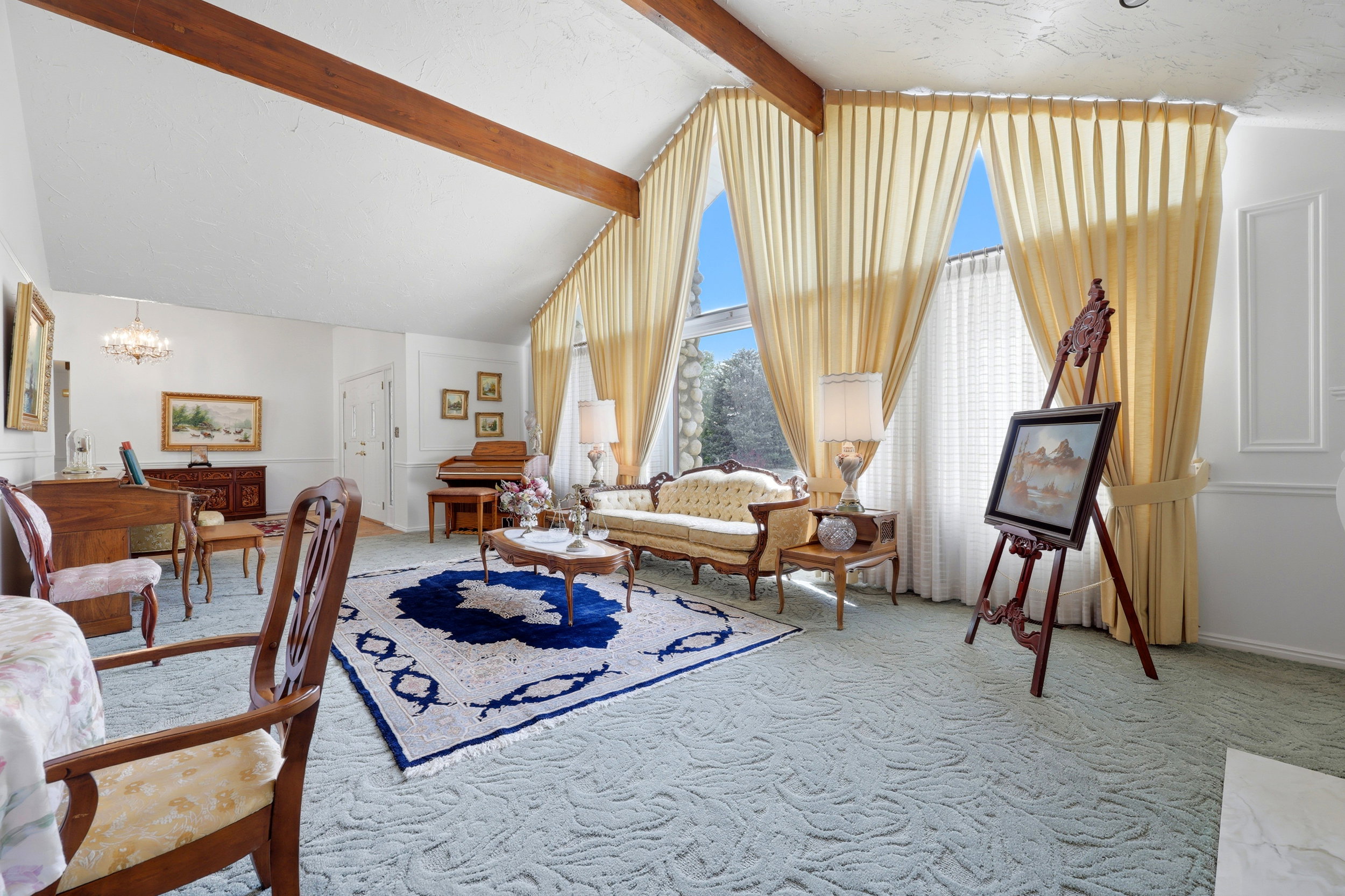 Spacious living area featuring a vaulted ceiling with exposed wood beams, expansive windows with floor-to-ceiling drapery, and textured carpeting