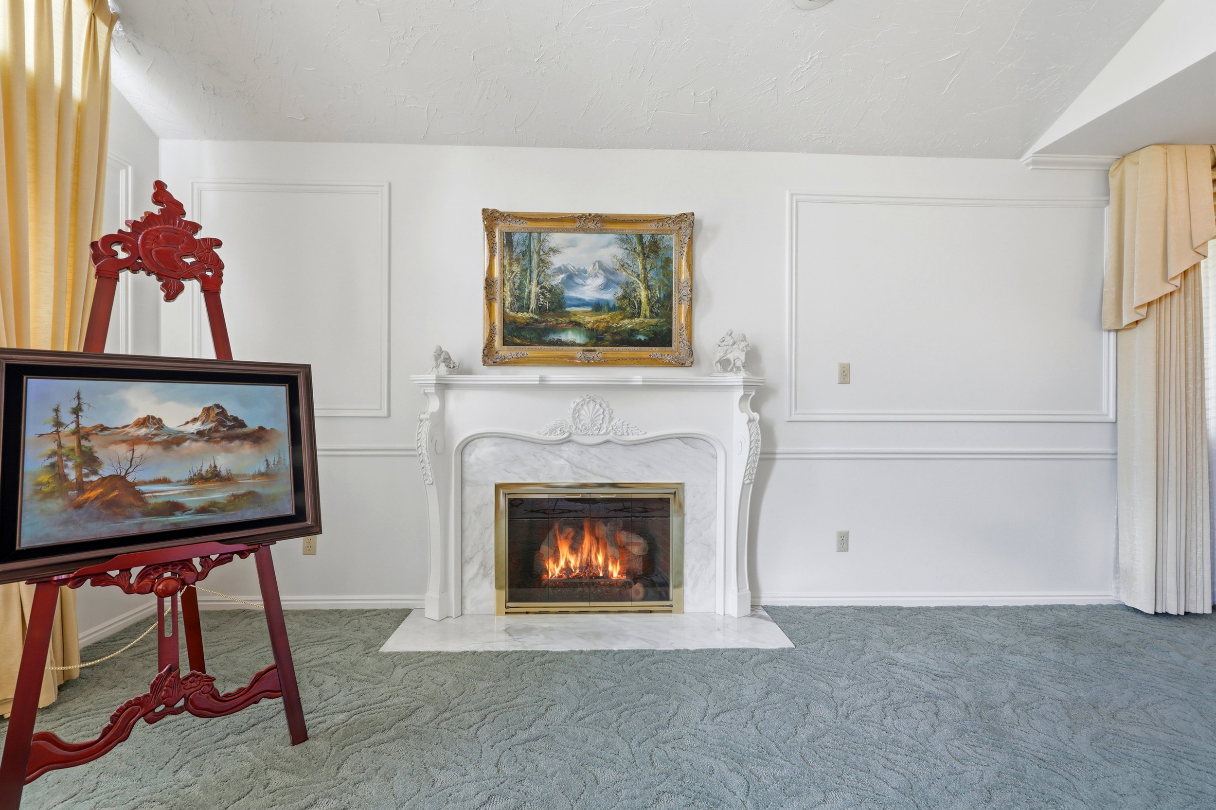 Ornate fireplace with a marble surround and hearth, framed by white paneled walls and light-colored drapes