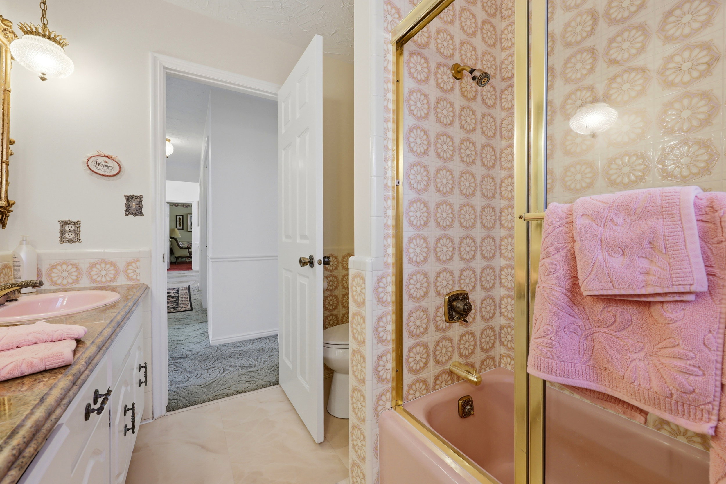Bathroom featuring a pink tub and coordinated wall tiles, a glass shower enclosure with brass-finish framing, a vanity with a pink sink, a stone-finish countertop, and white cabinetry with dark hardware