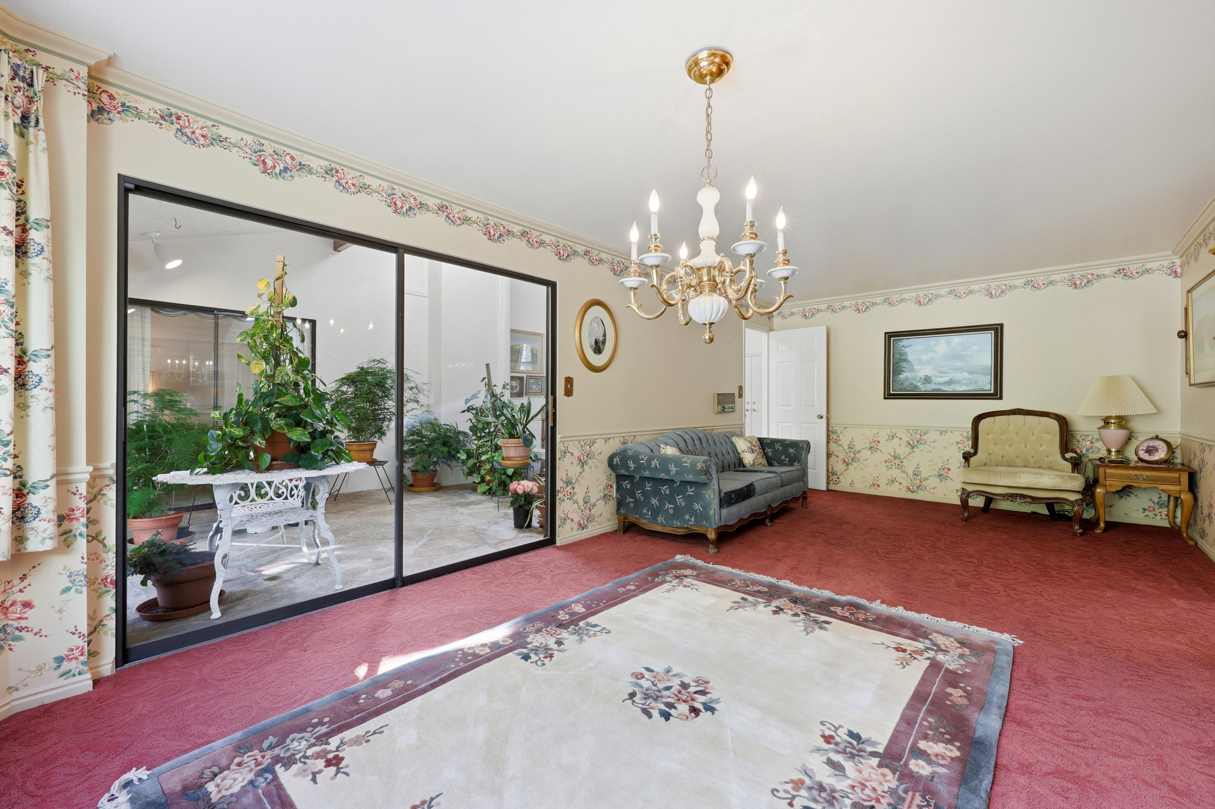 Spacious room with plush red carpeting, featuring an ornate chandelier and a large glass sliding door opening to an enclosed patio with various potted plants