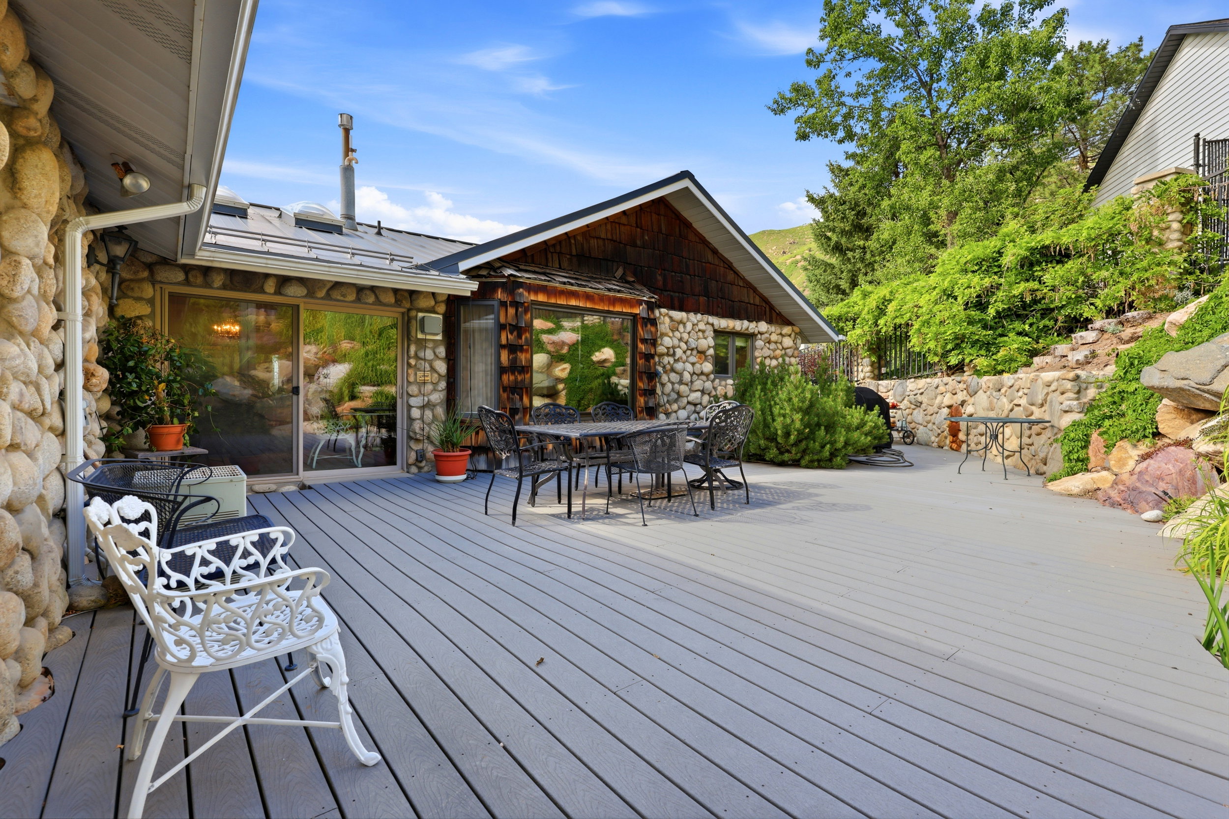 Expansive outdoor deck featuring a sliding glass door entry, stone veneer exterior, a stone retaining wall, and mature landscaping