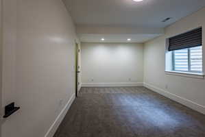 Spacious interior room featuring plush gray carpeting, recessed ceiling lighting, a window with a Roman shade, and white baseboards