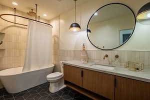 Bathroom featuring a freestanding tub with a ceiling-mounted showerhead, a dual-sink vanity with wood-finish cabinetry, a large round mirror, and geometric floor tiling