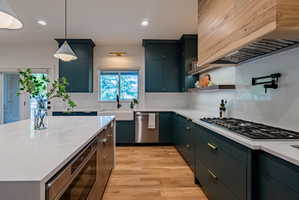 Gourmet kitchen featuring a central island with a white countertop, teal cabinetry, stainless steel appliances, a gas range with a pot filler, and wood-finish flooring