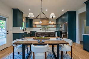 Open-concept kitchen featuring dark teal cabinetry, stainless steel appliances, a center island with seating, and wood-finish flooring