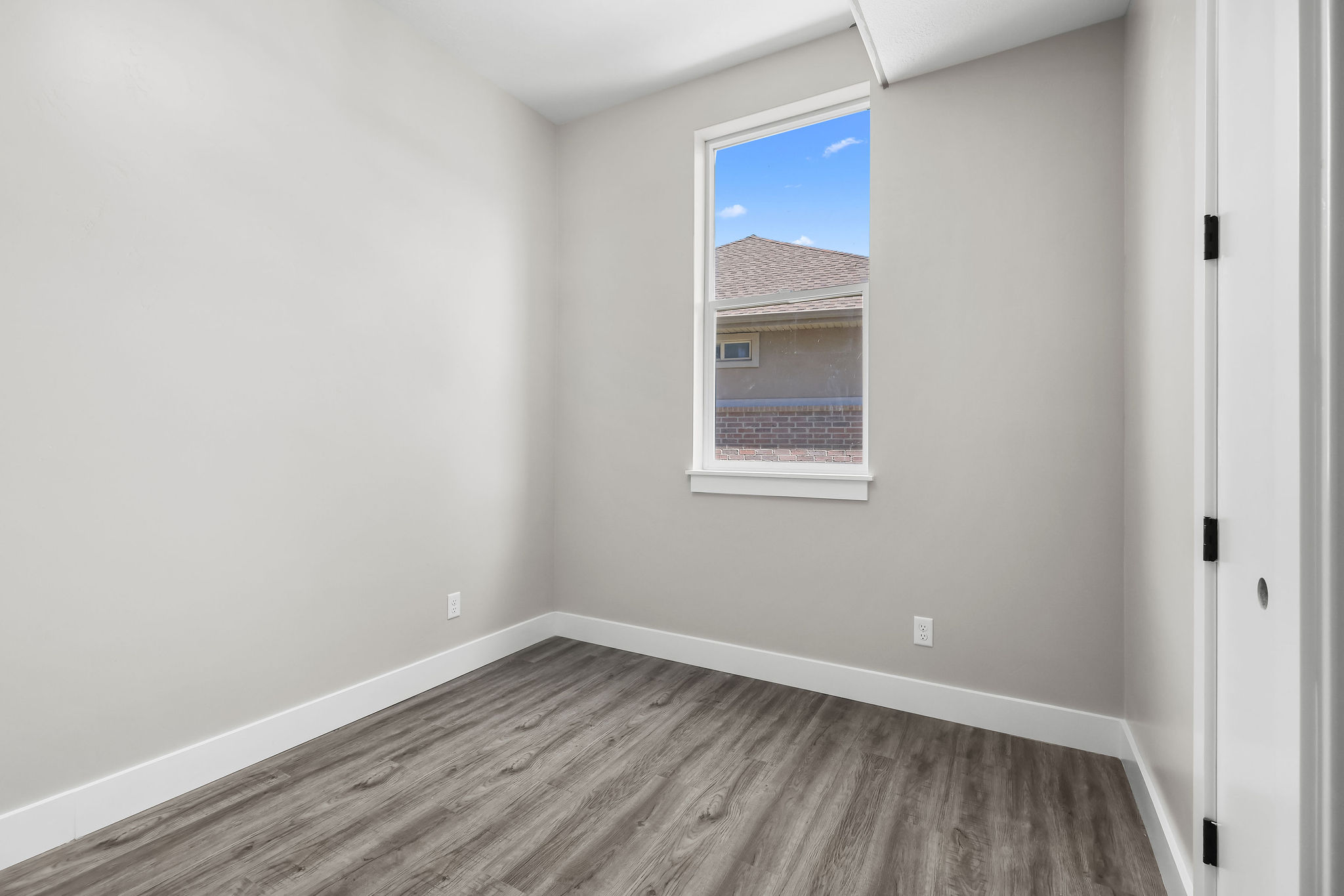 Light-filled room featuring a single window, wood-finish flooring, light grey walls, white baseboards, and a white door with black hardware