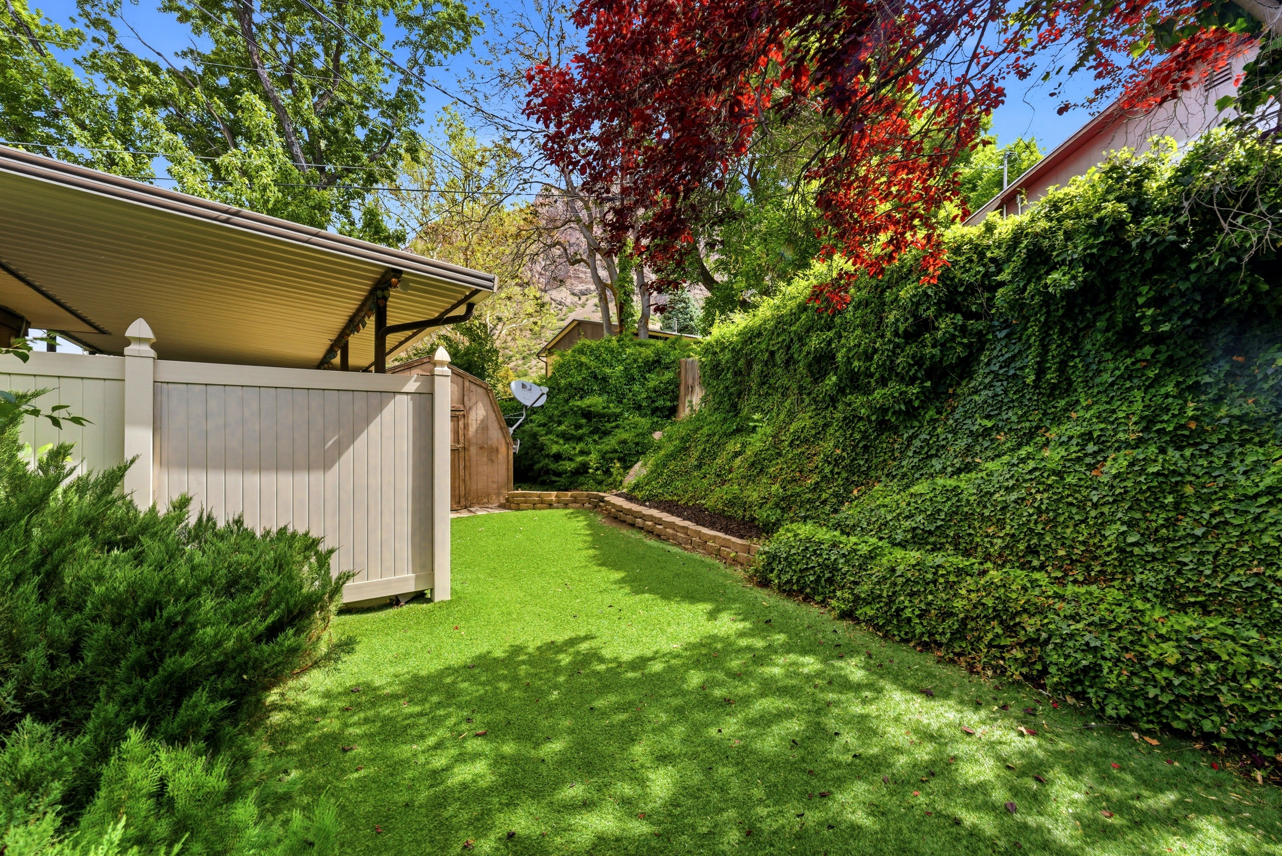 Fenced outdoor area featuring an artificial turf lawn, a sloped retaining wall with dense foliage, a privacy fence, and a covered patio structure