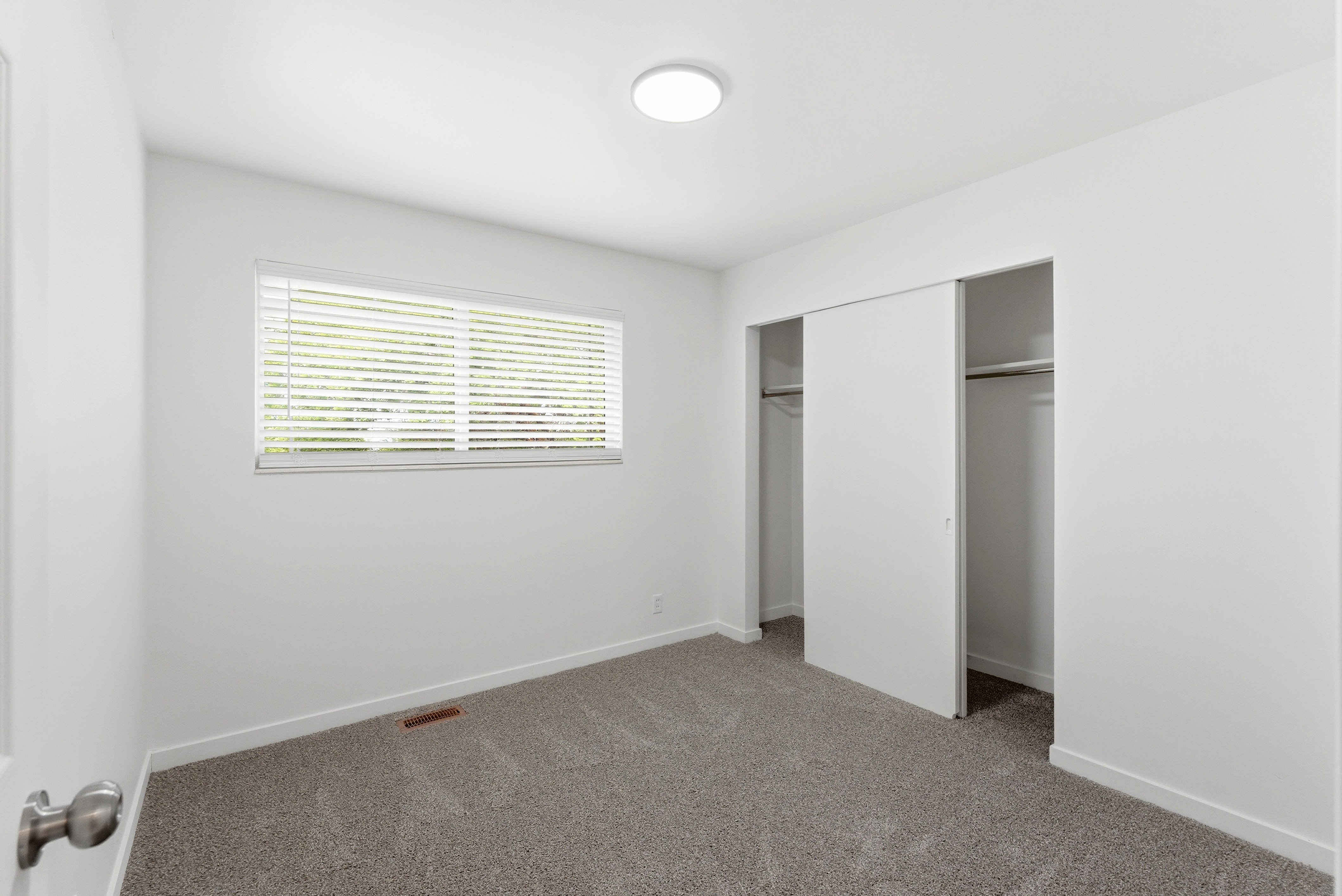 Room featuring light gray carpeting, crisp white walls, a single window with horizontal blinds, and a flush-mount ceiling light fixture