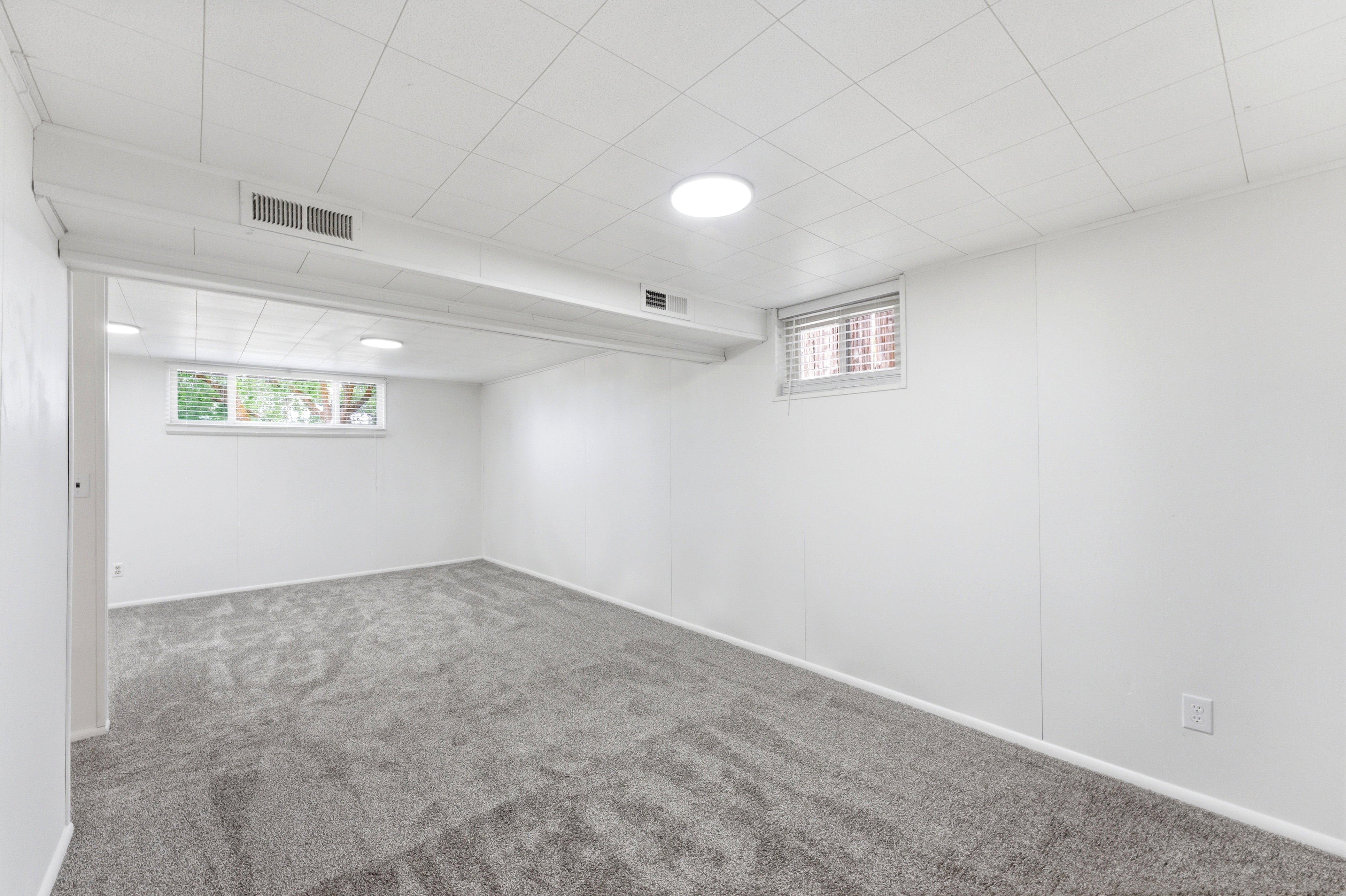 Spacious room featuring new gray carpeting, white painted walls, and a white drop ceiling with recessed lighting