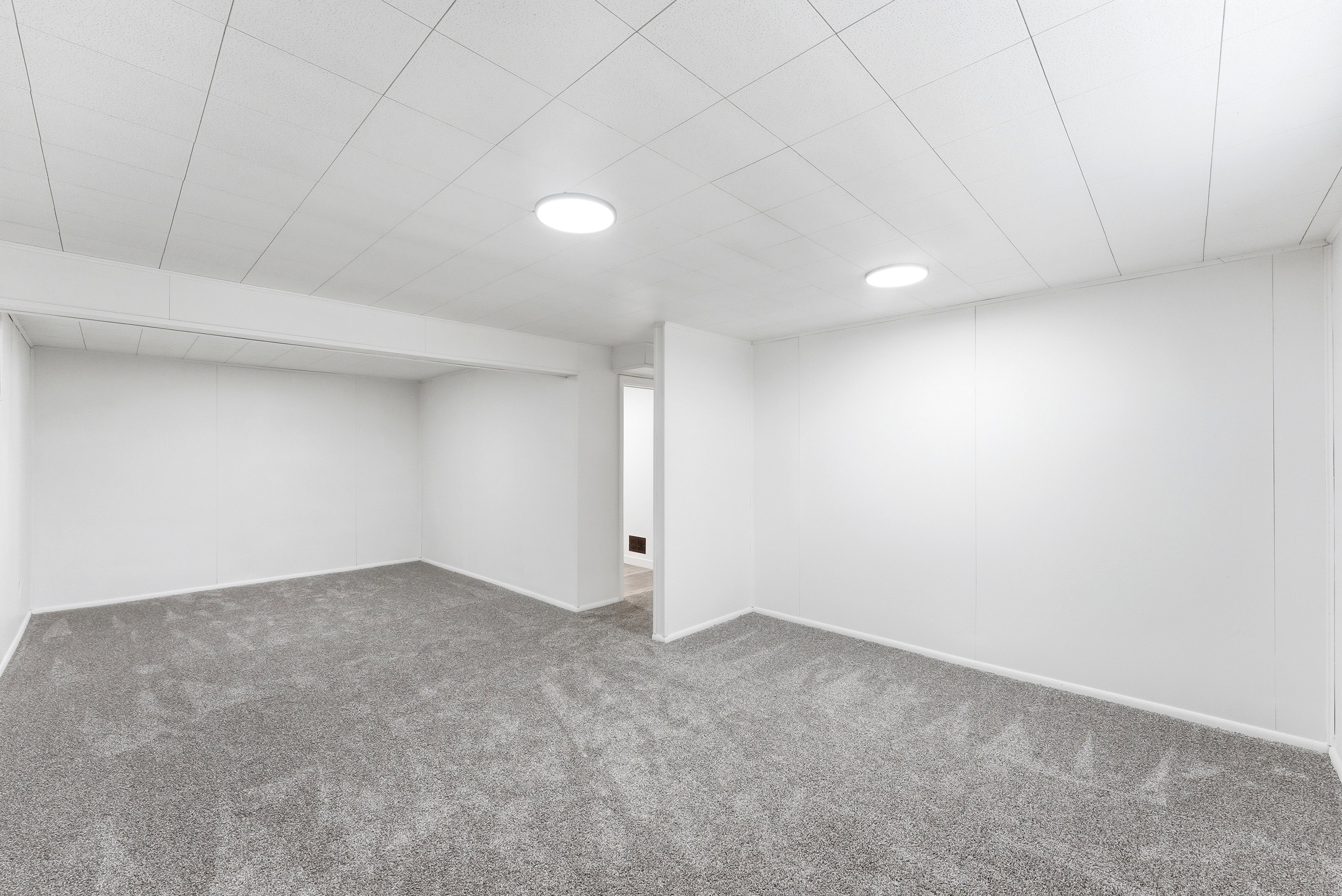 Spacious room featuring neutral carpeting, white walls, a drop ceiling with integrated circular lighting, and a visible hallway entrance