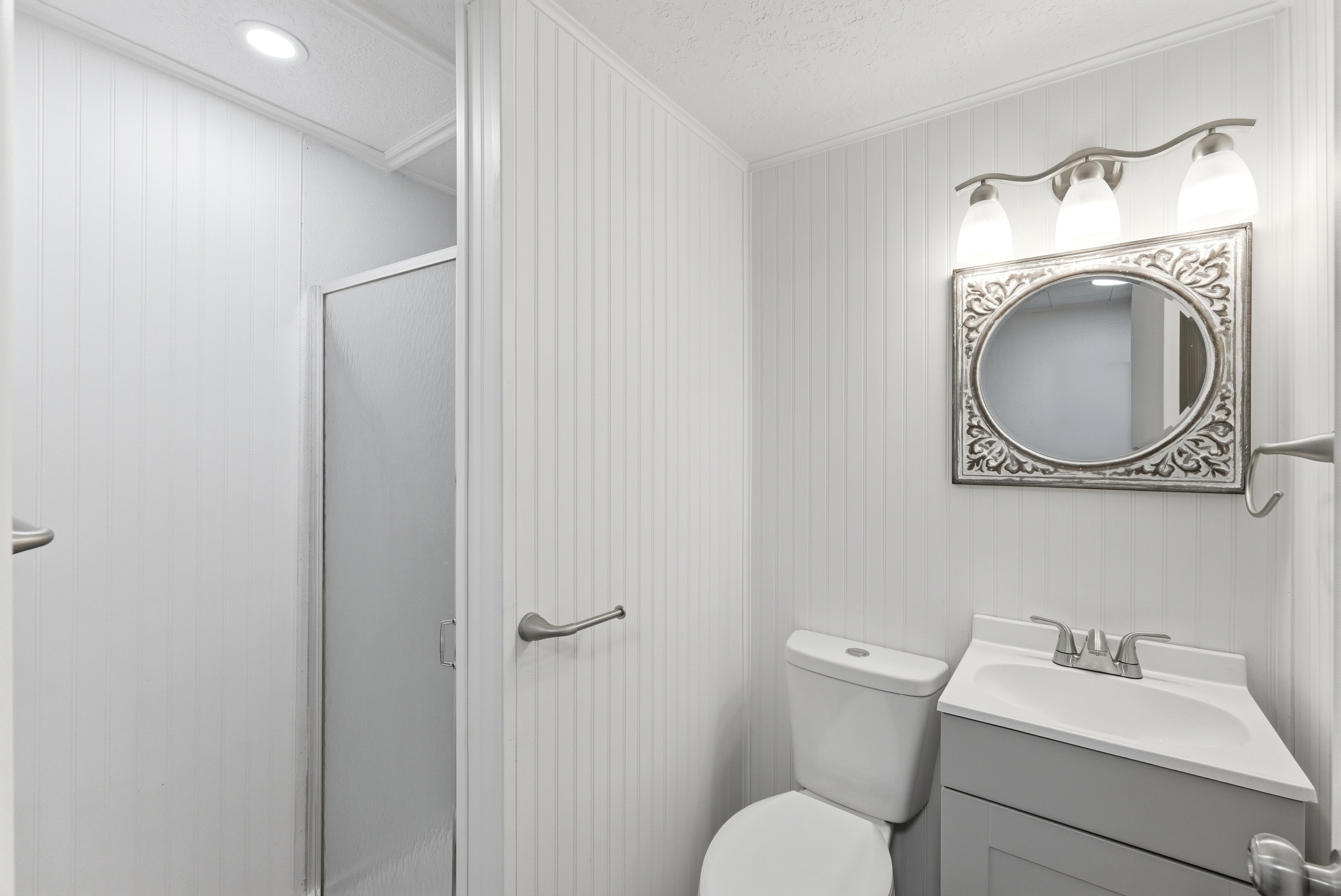 Bathroom featuring beadboard-style wall paneling, a corner vanity with a white countertop and gray cabinetry, a decorative framed mirror, and a three-light vanity fixture